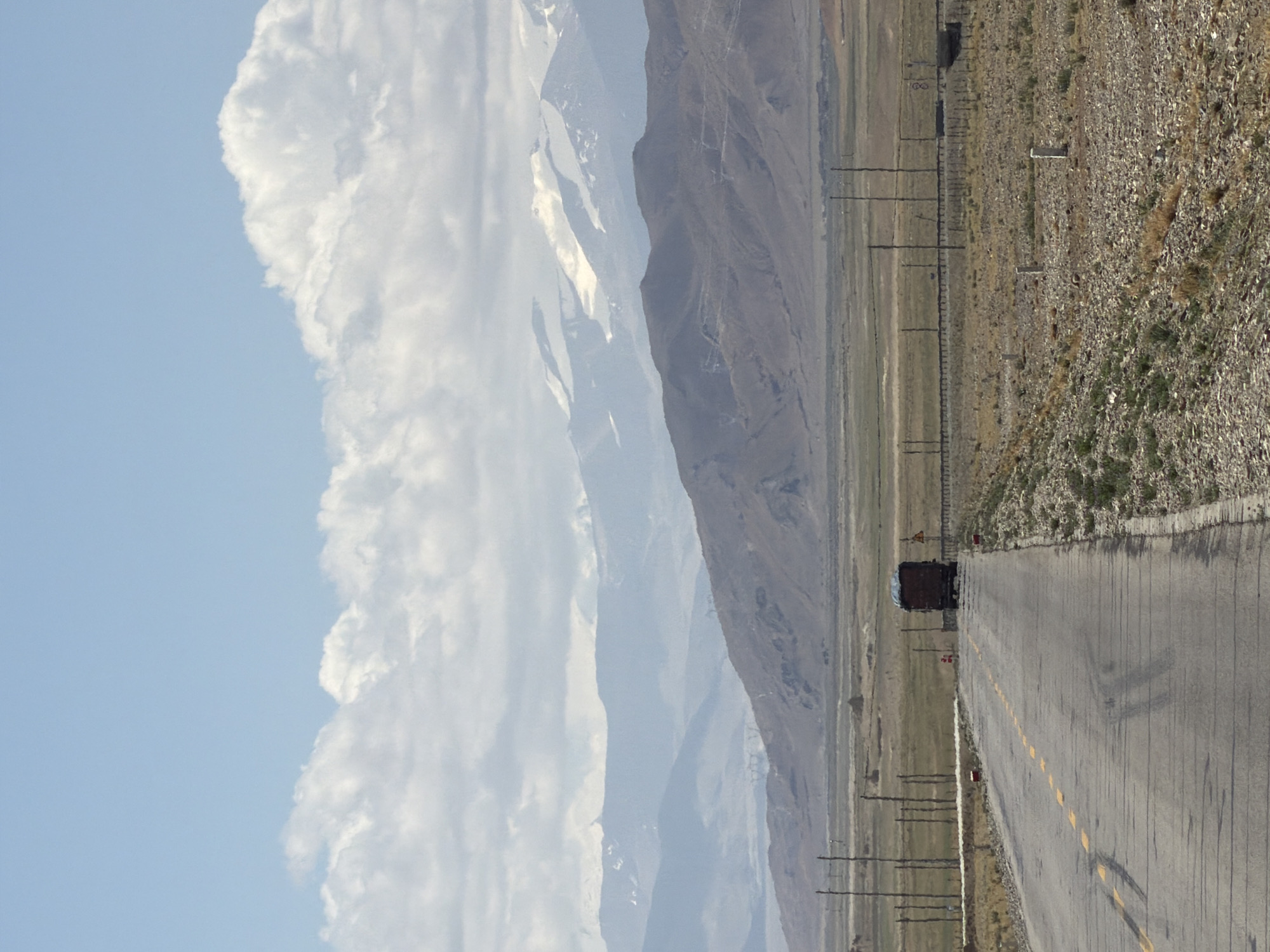 This image captures a vast, mountainous landscape traversed by a road, set in Mainpu township, China. The scene is depicted during daytime under a bright, partly cloudy sky.
In the foreground, a paved road, marked with a broken yellow centerline, stretches diagonally from the lower right towards the center distance. The road shows faint tire marks. To the right of the road, the ground is arid, stony, and sparsely covered with dry grass and scrub. A series of utility poles run parallel to the road on this side. Several small, rectangular white markers are visible along the roadside; one clearly displays the text "L 18", likely a kilometer or mile marker. A smaller, red-marked sign is also partially visible near a yellow diamond-shaped warning sign on the left side of the road, further down.
In the mid-ground, a dark, possibly black, truck is driving away from the viewer on the road, suggesting transport or travel through this remote region. To the left of the road, the terrain appears flatter and extends towards the base of the mountains.
The background is dominated by several layers of mountains. On the left, rugged peaks rise, some displaying patches of white, possibly snow or lighter rock formations, indicative of high altitude. These mountains appear hazy and blueish in the distance due to atmospheric perspective. The sky above is a pale blue, adorned with large, voluminous white cumulus clouds that fill a significant portion of the upper left. The overall impression is one of isolation and a journey through a grand, natural environment.