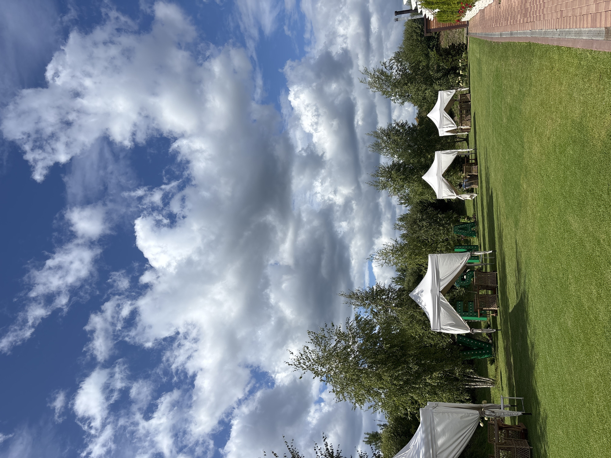 The image depicts an outdoor scene on a grassy lawn in Pokrovskoe, Russia, under a sky filled with dramatic clouds. Several white canopies, likely for outdoor dining or events, are set up on the lawn. Each canopy covers a seating area with wicker furniture, including chairs and tables. In the background, a line of trees forms a natural border, with some taller birch trees interspersed with other greenery. To the right of the lawn, a brick pathway runs alongside a building with white vertical accents, possibly part of a restaurant or event venue. The overall impression is of a well-maintained outdoor space prepared for patrons.