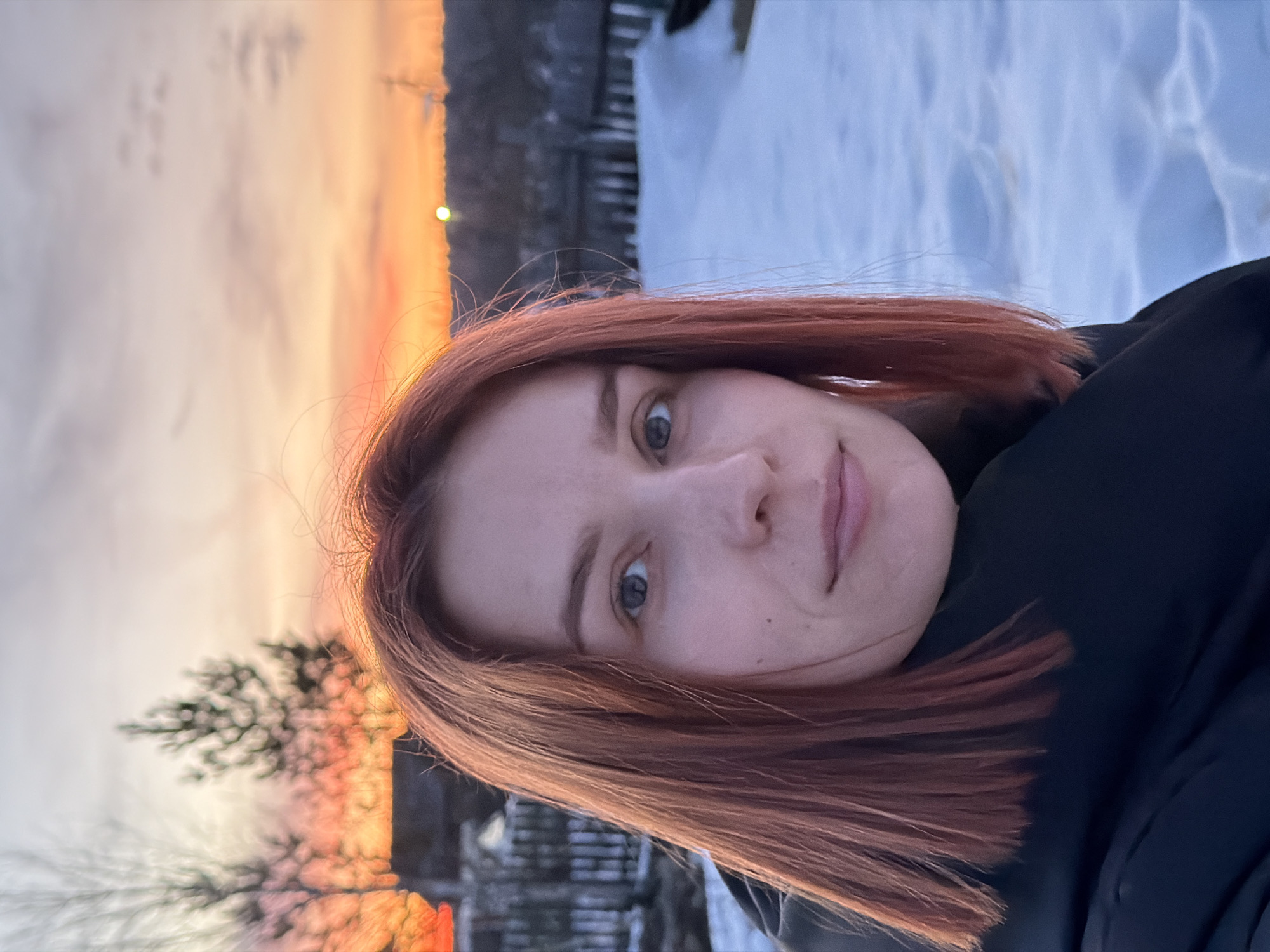 A woman with reddish-brown hair is the subject of this selfie taken outdoors. She has blue eyes and a fair complexion. She is wearing a black garment. The background reveals a snowy environment and what appears to be a vibrant sunset or sunrise, with hues of orange, yellow, and gray. There are silhouettes of trees and fence-like structures visible in the background. Based on the prompt, the location is implied to be Chaikovsky, Russia, though this is unconfirmed from the image alone.