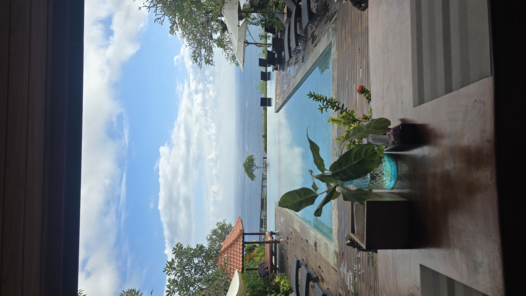 The image shows a view from a partially shaded indoor space looking out toward the ocean, likely from a hotel or resort in Kuta Selatan, Indonesia. A small infinity pool is visible, designed to give the illusion of blending into the sea. Several lounge chairs are positioned near the pool's edge.
The ocean extends to the horizon, and the sky is bright blue with scattered clouds. A small tree sits at what seems to be the edge of the cliff where the infinity pool resides.
In the foreground, there is a wooden table with a small potted plant. The edge of a window or doorway frames the view.