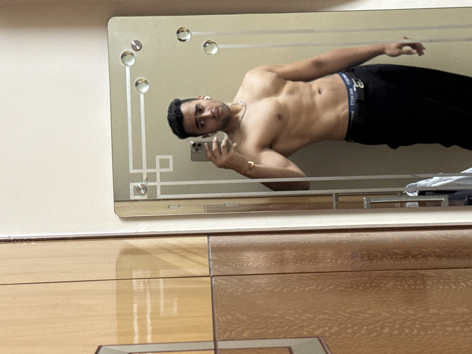 A shirtless man in black pants takes a selfie in a decorative mirror. He wears a gold bracelet and a necklace, with earbuds in his left ear. The mirror reflects him from the waist up, revealing a toned physique. The mirror has a beige background with etched designs and decorative circles. It's mounted on a wall above a glossy wooden surface with a geometric pattern.