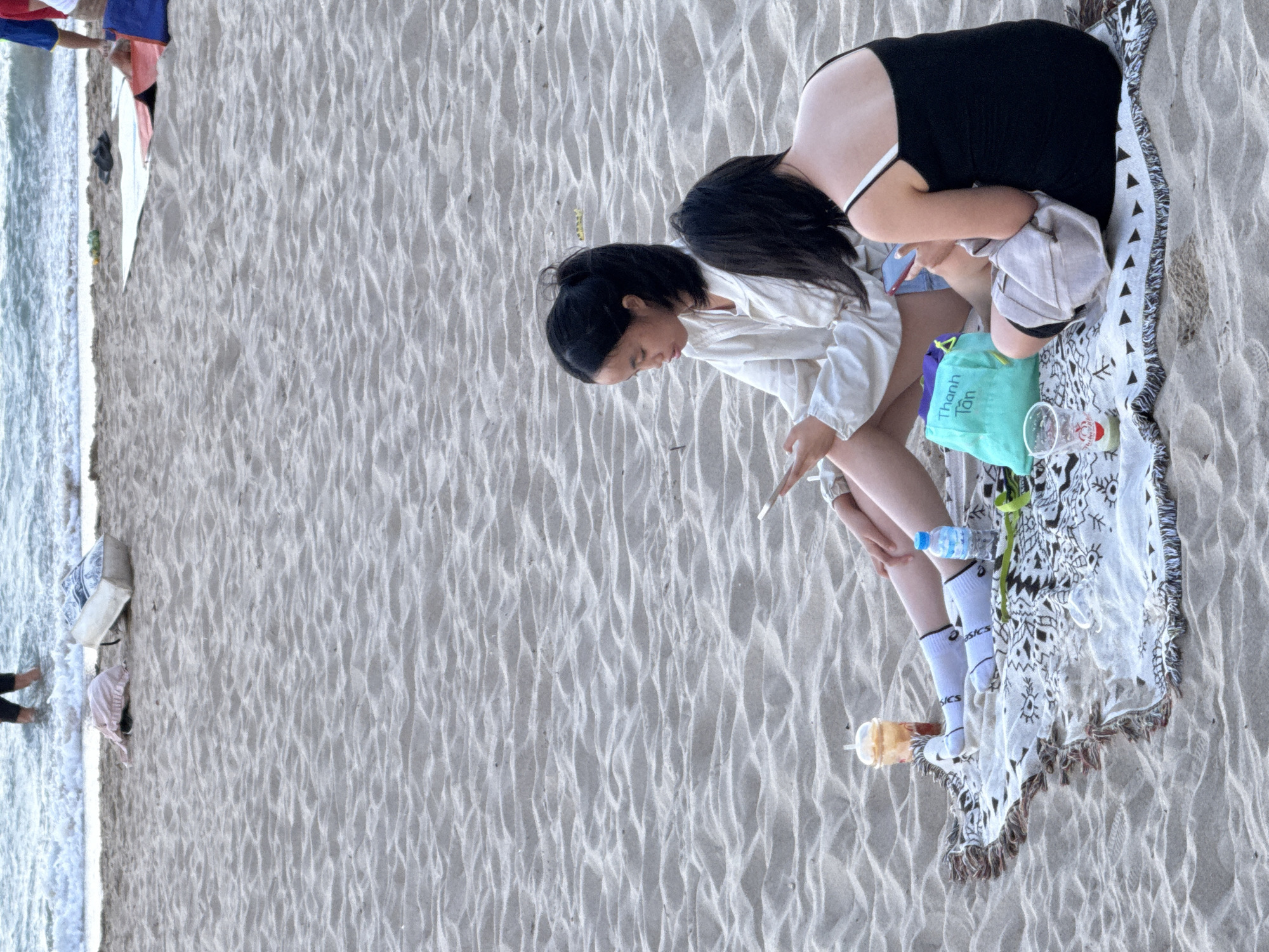 Two young Asian females are seated on a patterned blanket on a sandy beach. The person on the left, facing right, wears a white long-sleeved shirt and white socks. She is holding a smartphone in her right hand and looking down at it. Next to her are a plastic cup with a straw and a bottle of water. The person on the right, facing left, wears a black slip dress and is also holding a smartphone. In front of them is a turquoise bag with Vietnamese writing and a plastic cup. The background shows gentle waves of the ocean and sand. The scene is likely the coast of Nha Trang Ward, Vietnam, given the context provided.