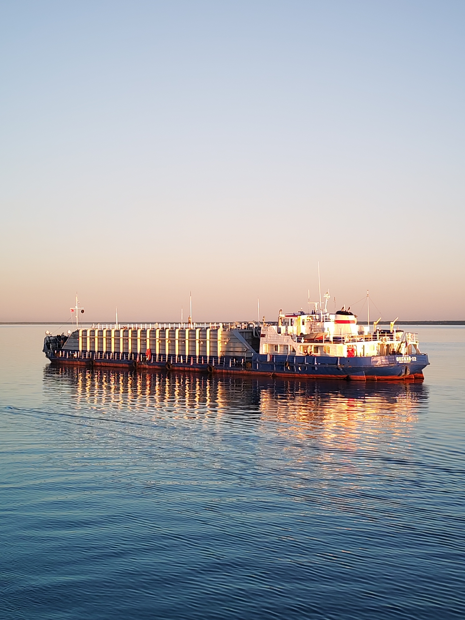 A wide shot depicts a large, dark blue vessel navigating calm, deep blue water under a clear, gradient sky. The sky transitions from light blue at the zenith to a pale orange-peach hue near the horizon. The vessel, identified as "ОКСКИЙ-22" on its stern, is oriented with its bow towards the left, displaying a long, flat deck area adorned with numerous vertical, light-colored stanchions that are strongly illuminated by low-angle, golden sunlight. The stern section features a multi-level white superstructure with windows, a bridge area, and a red-and-white striped funnel. The sun's light originates from the left, casting a warm, golden glow on the vessel's starboard side and its vertical structures. This illumination is vividly reflected in the rippling water below the vessel, creating elongated, wavy patterns of golden light and dark blue. A subtle wake trails behind the stern, indicating movement. A thin, dark line on the distant horizon suggests a landmass or distant shore. No individuals are visibly present on the vessel or in the surrounding environment. The scene is located in Unknown, Unknown.