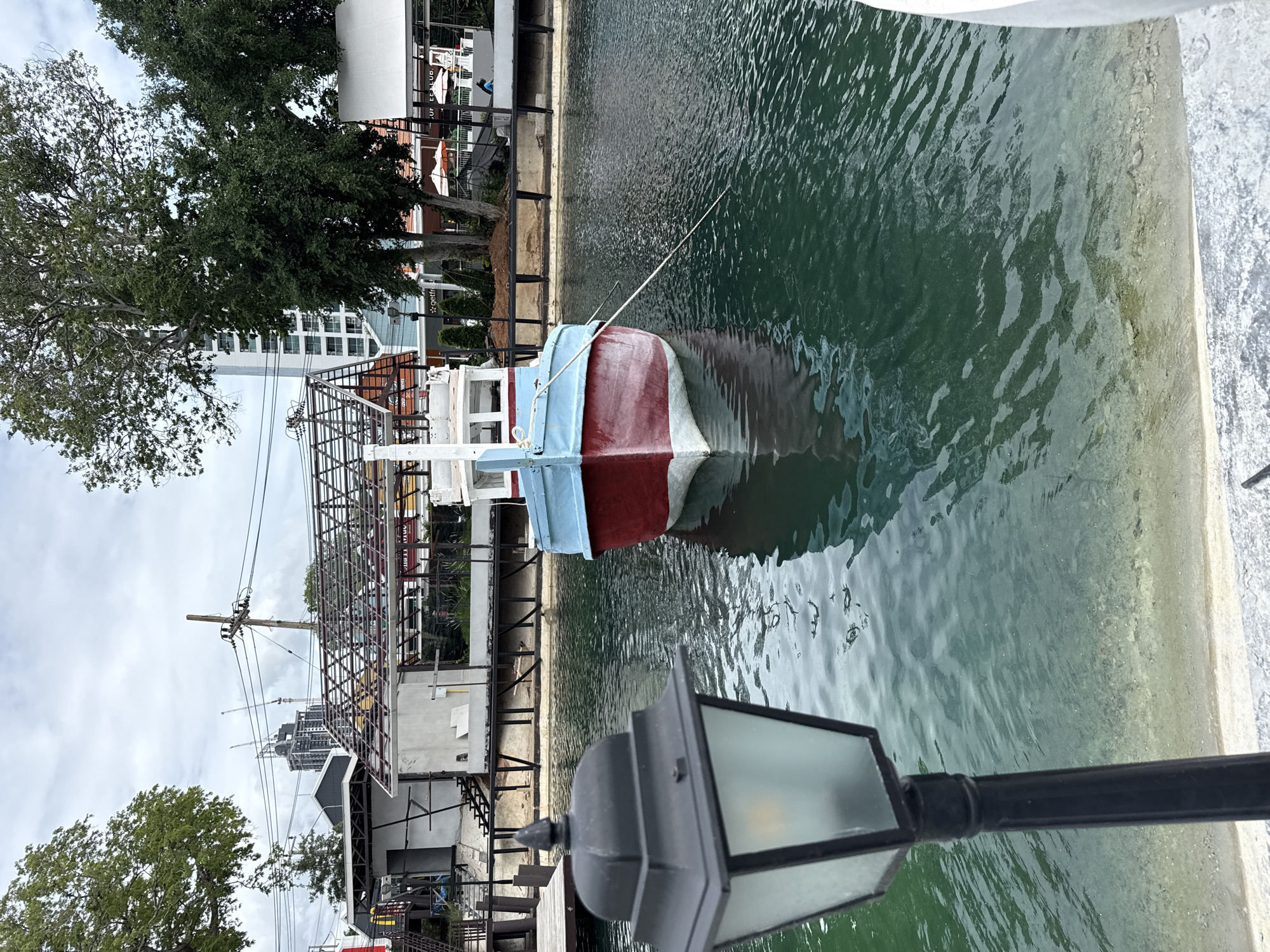 In Pattaya City, Thailand, a red and blue boat is moored in a body of water. A black lamppost with a light is in the foreground, to the left. In the background, there are buildings and trees. The sky is overcast.