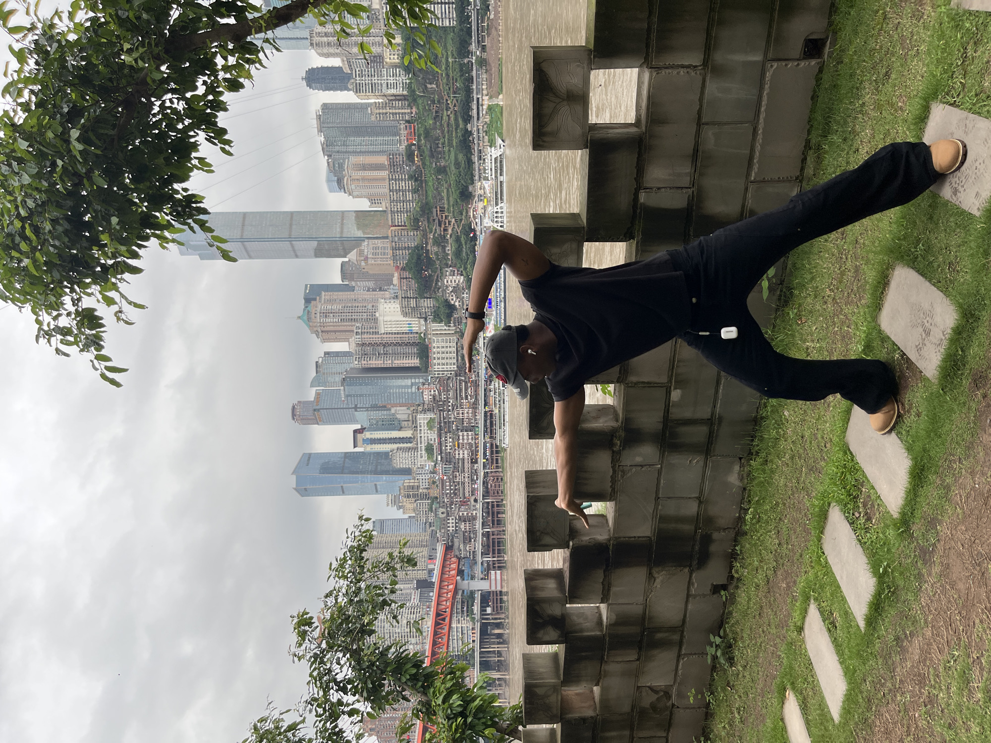 A man in black clothing and tan shoes strikes a pose on a stone structure next to a grassy area. He's wearing a gray baseball cap and appears to be interacting with the structure, placing his hand on it. In the background is a dense cityscape under a cloudy sky, featuring numerous high-rise buildings. The city visible is Yuzhong District, China, as identifiable by the architecture. There are trees in the foreground. The photograph appears to be taken at an unusual angle, rotated counter-clockwise.