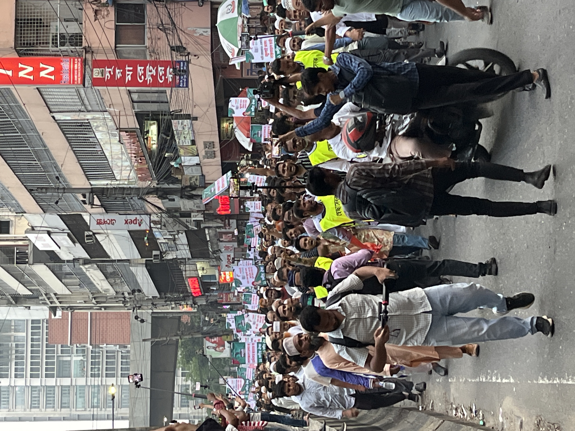 A large crowd of people are marching in a street in Dhaka, Bangladesh. Many are holding signs, some with writing in Bengali. Buildings line the street, with visible signs and banners, one of which reads "Apex" in English. Some people are taking photos and videos with their phones. A person on a motorcycle appears to be filming or photographing the crowd. The scene appears to be a demonstration or protest.