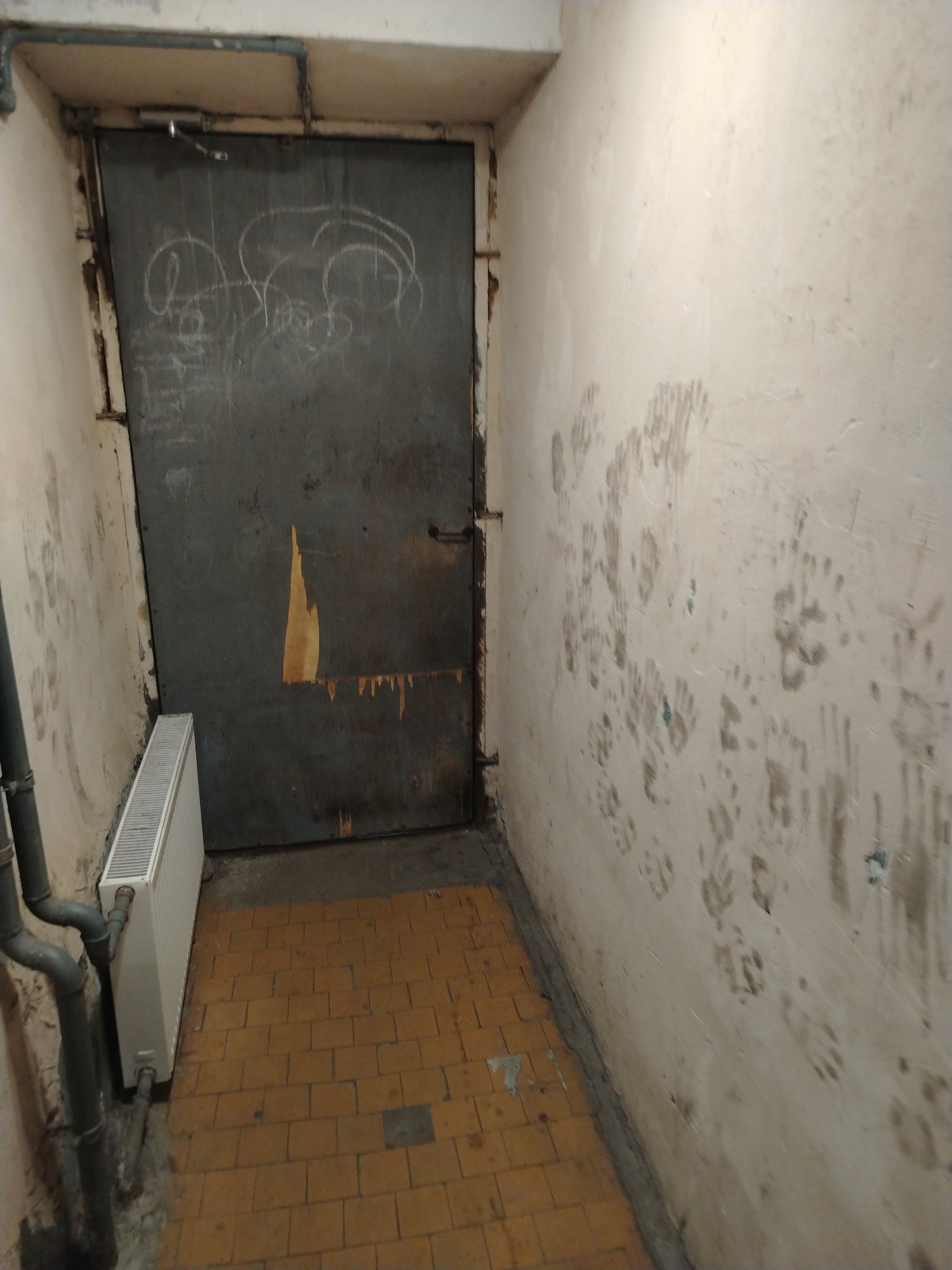 This is a photograph of a hallway with a metal door and a radiator. The walls are plastered and marked with numerous handprints. The floor is tiled with yellow square tiles. The metal door appears to be an exterior door, with a handle and hinges visible. There are some markings or graffiti on the door. The radiator is white and positioned to the left of the door. The hallway appears to be in a utilitarian or industrial setting, possibly a basement or service area.
Given that the Vyborg Castle is a prominent landmark in Vyborg, Russia, and is known for its historical significance and various interconnected buildings, it is plausible that this hallway could be located within the castle complex or in an associated structure in the city of Vyborg. The presence of a metal door and radiator suggests a functional rather than decorative space, which might be found in the less frequented or utility areas of such a historical site. The handprints on the wall could indicate a place where people have historically gathered or passed through, or they could be more recent markings.
