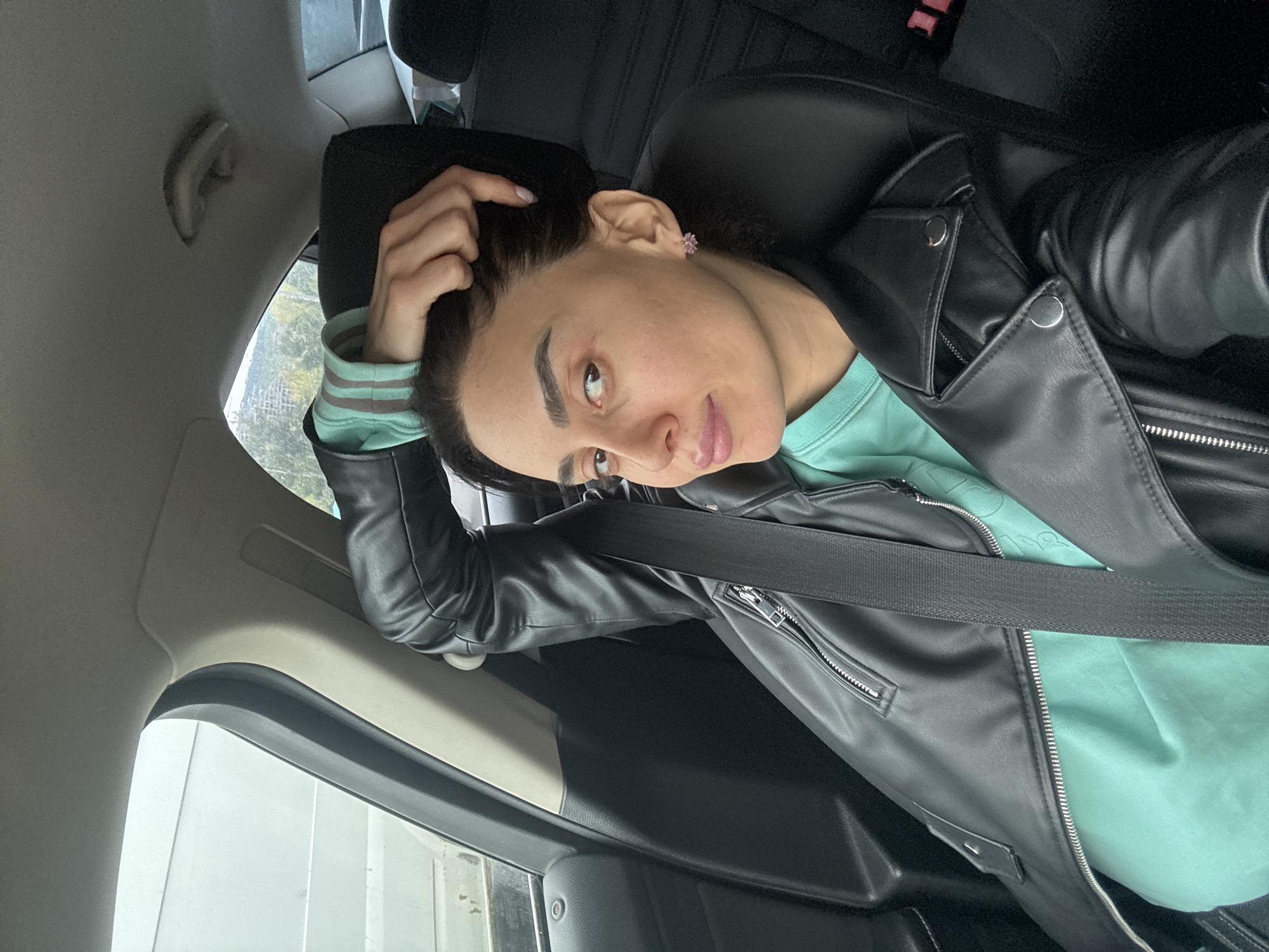 A young woman is captured in a sideways-oriented photograph, seated in the back of a car during what appears to be daytime in Tbilisi, Georgia. She has dark hair and looks directly at the camera with a neutral, relaxed expression, her left hand gently resting near her head. She wears a small, light pink, flower-shaped stud earring in her visible left ear.
Her attire consists of a black faux leather jacket with silver zippers and snap buttons, worn over a light teal or mint green long-sleeved top. A black seatbelt is visible across her chest. The car's interior features black leather seating and grey trim panels, with a headrest behind the woman. Through the car windows, a blurred outdoor background suggests trees or distant structures, indicating the vehicle is either parked or moving slowly. There is no visible text in the image.