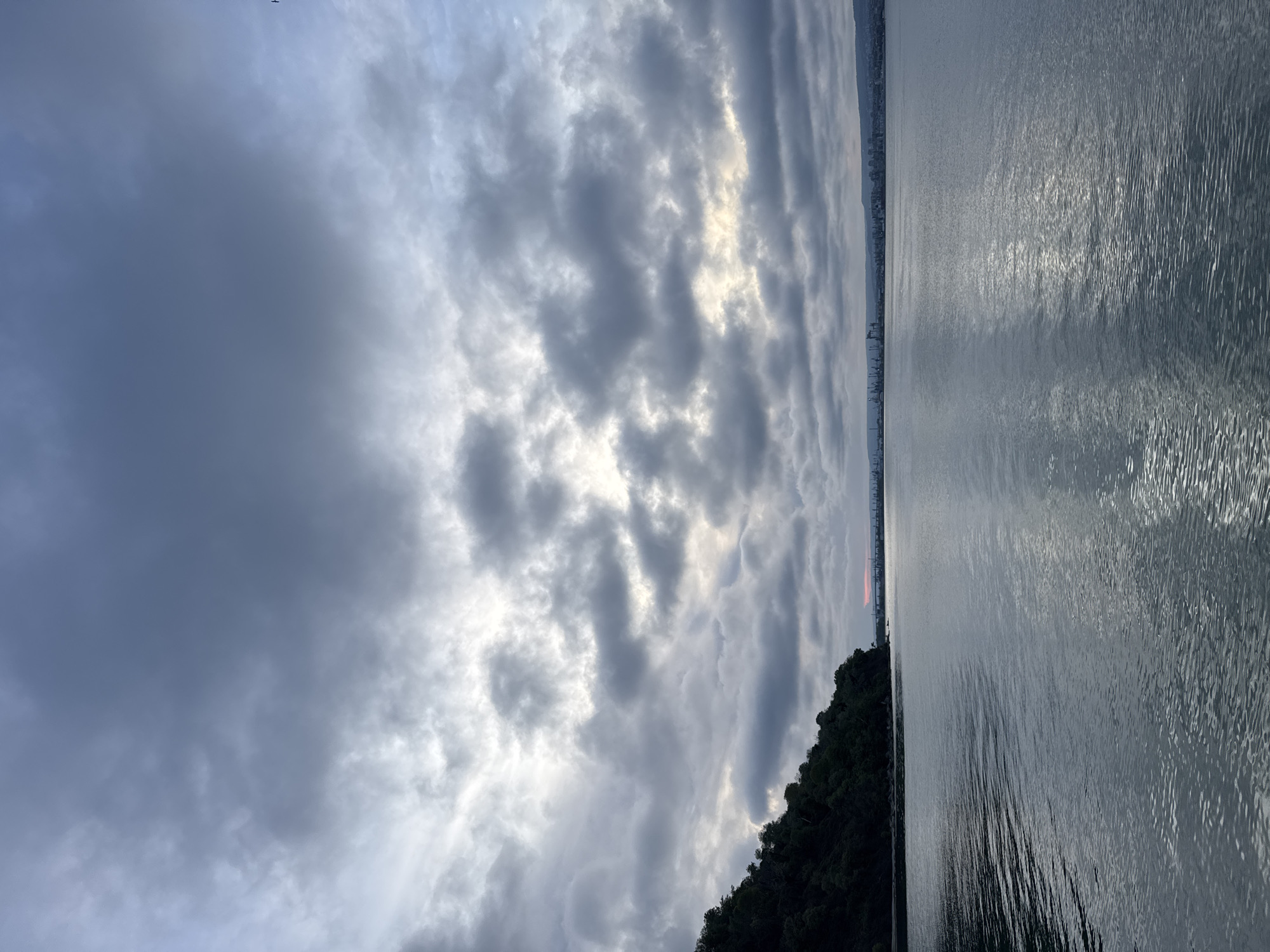 The image shows a body of water, likely the Black Sea, under a cloudy sky. The clouds are predominantly gray with patches of lighter tones where sunlight is breaking through. The water is textured with ripples, reflecting the light from the sky. A dark green, possibly forested, shoreline is visible on the lower right side of the image. A distant urban skyline stretches along the horizon. Given the context of Varna, Bulgaria, the location is likely a coastal area near the city. There are no people or direct human activities visible. The scene is dominated by natural elements.