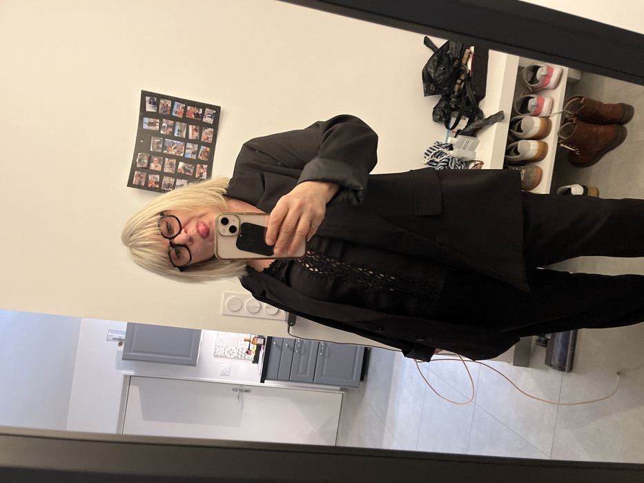 A woman with blonde hair and black glasses poses for a mirror selfie. She wears a black blazer and a top with lace detailing. A black-framed picture collage hangs on the white wall behind her. To her right, a shoe rack holds various shoes and bags. A glimpse of a kitchen with gray cabinets is visible in the background, along with light gray flooring.