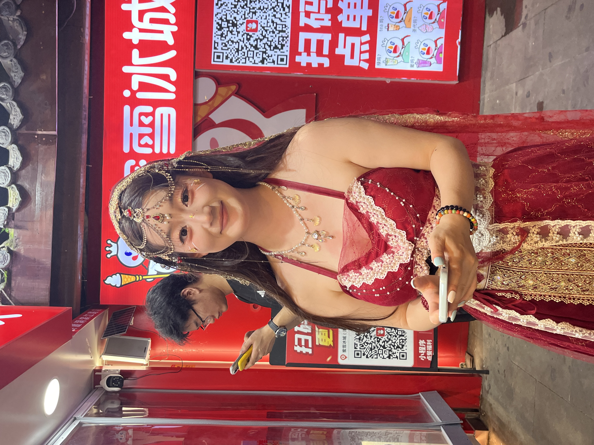 In Yuzhong District, China, a young woman in a red, ornate dress, adorned with jewelry and face paint, stands holding a smartphone. She is positioned in front of a bright red storefront featuring Chinese characters and QR codes. Behind her, a man in glasses looks at his phone. The environment appears to be an outdoor market or commercial area, indicated by the storefront and the pavement. The woman's attire suggests a cultural performance or event.