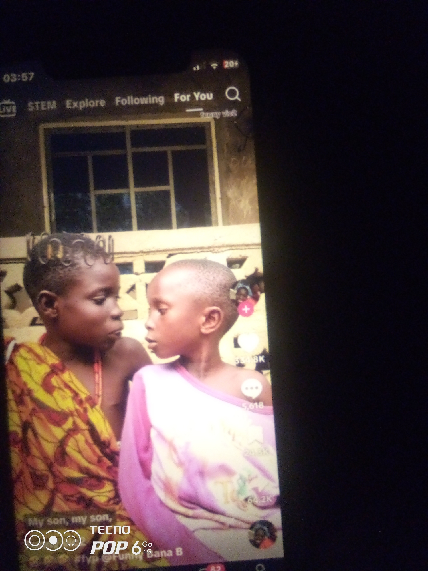 A vertical, slightly blurry, close-up, eye-level shot shows two young children facing each other on a mobile phone screen. The phone appears to be a TECNO POP 6, indicated by text at the bottom of the screen. The children are dark-skinned with short hair. The child on the left wears a yellow and red patterned garment and a beaded necklace. The child on the right wears a pink and white tie-dye shirt. They are looking into each other's eyes with serious expressions. The background is indistinct but appears to be a wall with a window or grate. The phone screen displays interface elements typical of a social media or video-sharing app, including the time "03:57", navigation tabs "LIVE STEM Explore Following For You", a search icon, and numerical engagement metrics like "334.8K" and "64.2K". The text "My son, my son," is visible below the children. The overall impression is of an intimate moment captured on a digital device. There is no information in the image to indicate the city of Howayi, Nigeria.