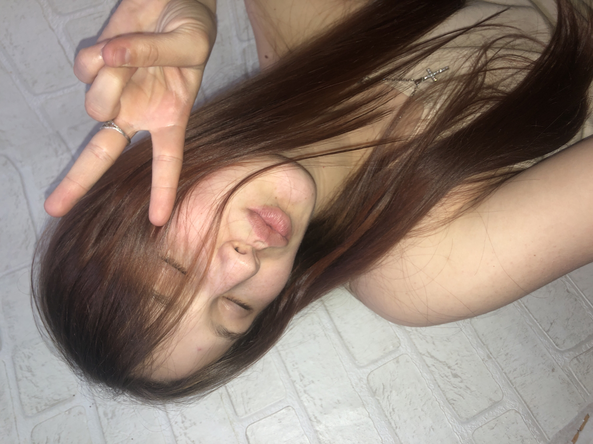 A woman is lying on a white tiled floor. She is positioned diagonally in the frame, with her head tilted towards the left. Her long brown hair is partially covering her face. She is making a "peace sign" with her left hand. She is wearing a ring on her left ring finger. The woman has her eyes closed. She is wearing a cross necklace. The location appears to be indoors, but it is impossible to determine if the location is in the city of Фоки, Russia.