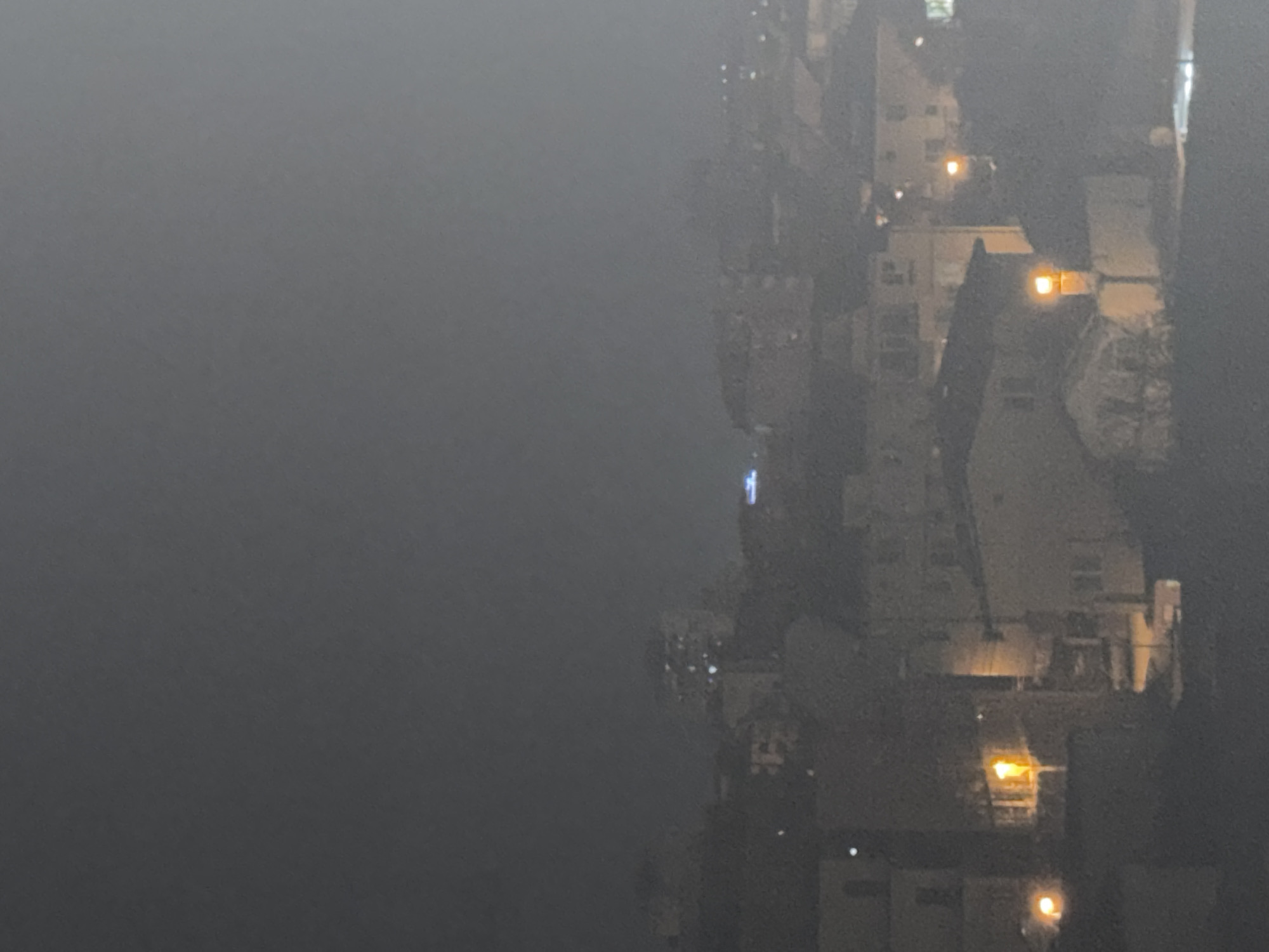 The image shows a view in Varna, Bulgaria, possibly taken from a high vantage point. The visibility is severely limited, with a dense fog or smog obscuring much of the city. Structures, possibly buildings, are dimly visible on the right side of the image. Their shapes and details are hard to discern, but some appear to have lights, which emit a yellow or orange glow. The left side is mostly obscured by the fog. The image suggests conditions of low visibility and possibly air pollution in an urban environment.