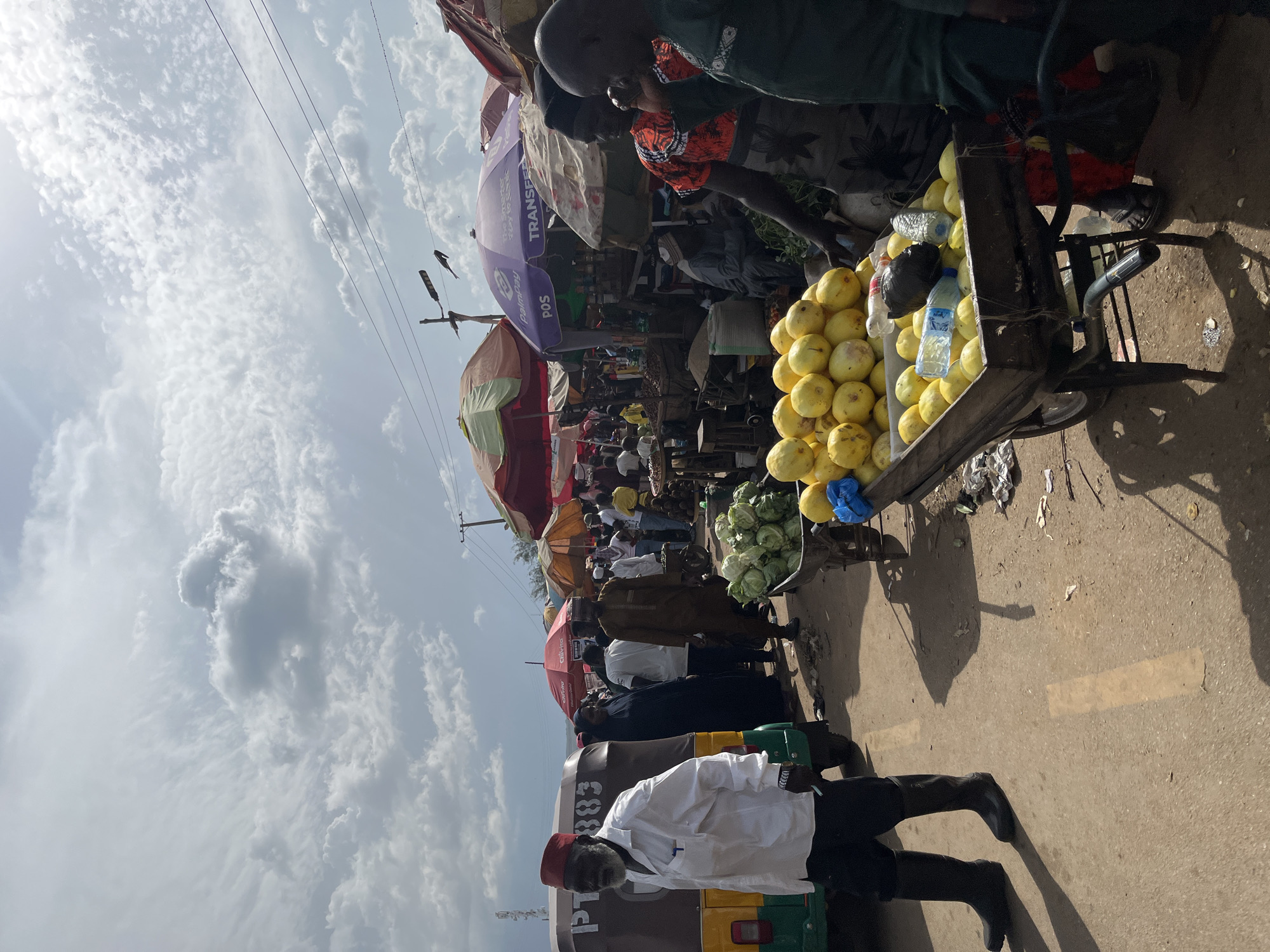 The image depicts an outdoor market scene in Rafin Par, Nigeria, under a cloudy sky. In the foreground, a man wearing a white shirt, black trousers, and tall black boots walks towards the left. He has a red cap on his head. To his right, a wooden cart is filled with yellow fruits, possibly lemons or mangoes, with several plastic bottles and a black object on top. Behind this cart, another cart holds cabbages.
In the mid-ground, a bustling market is visible with numerous people, stalls, and umbrellas providing shade. Several individuals are seen interacting, some appearing to be vendors and customers. A three-wheeled vehicle, possibly an auto-rickshaw, is parked on the left side of the image. Various market goods are displayed, including what appear to be textiles and more produce. The ground is paved with dirt and some debris is scattered around. In the background, electrical wires and a utility pole are visible against the sky. The overall atmosphere suggests a busy commercial hub.