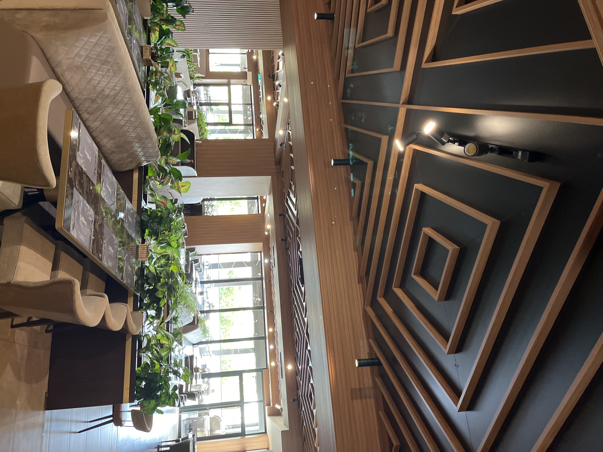 The image shows the interior of a modern restaurant in Shumen, Bulgaria. The ceiling features a complex geometric design with wooden beams against a dark background, and several track lights are installed. The restaurant has a contemporary aesthetic with comfortable seating, including a long upholstered booth and individual chairs. Tables are set with menus, and potted plants are used as decorative elements, particularly along the banquette seating. Large windows offer a view of an outdoor area or patio. The overall atmosphere suggests a stylish and well-maintained dining establishment.