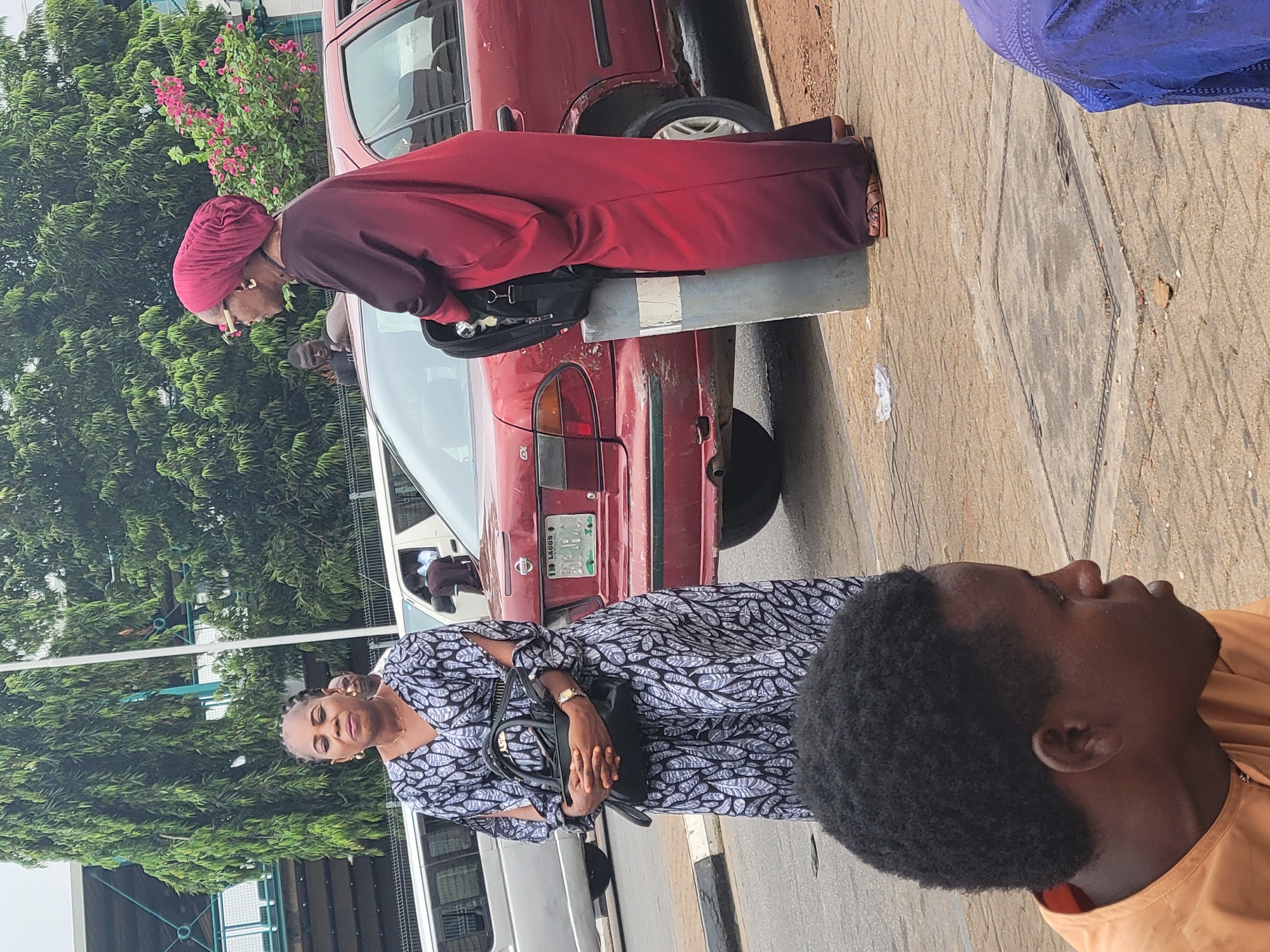 The image shows three individuals standing near a red car on a street. The license plate on the car reads "LAGOS, BUQ-18764". A woman in a dark and white patterned dress stands with her hands over a black handbag. Behind her, another woman in a maroon robe and turban leans over the car. A man with short black hair is visible in the foreground wearing an orange shirt. The street has sidewalks. Trees are visible in the background. The photo may be taken in Abuja, Nigeria.
