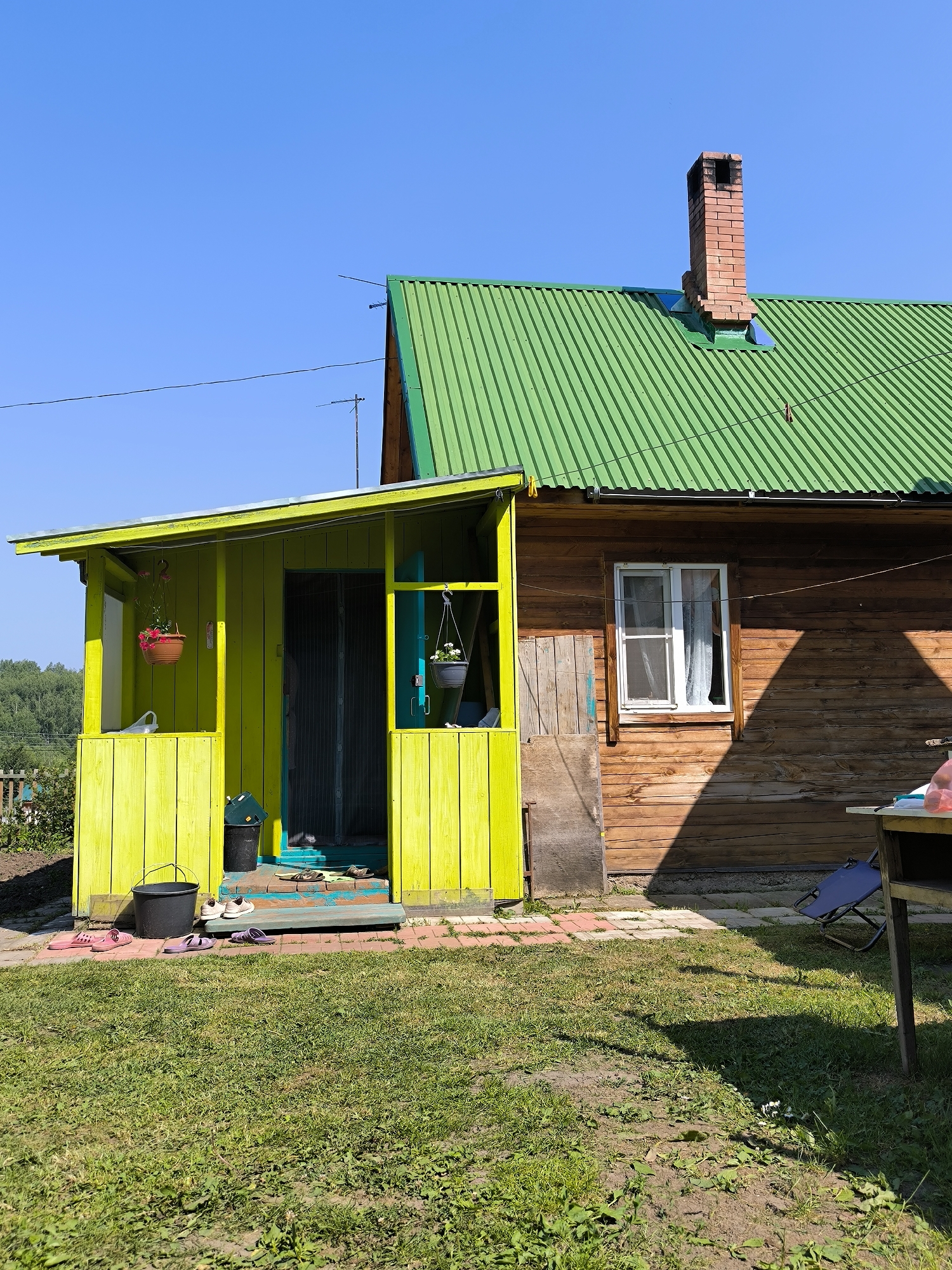 The image depicts a wooden house with a bright green metal roof and a contrasting bright yellow porch in the village of Maiskii, Russia. The house is constructed of horizontal wooden planks, with a single white-framed window visible on the right side, featuring white curtains. A red brick chimney stands on the peak of the roof. The porch is painted a vivid yellow and has vertical wooden planks. Several pairs of footwear, including sandals and closed-toe shoes, are placed on the porch steps and the adjacent ground. A black bucket and a dark green trash bin are situated on the porch. Two hanging flower baskets, one with red flowers and another with white flowers, are suspended from the porch roof. The foreground is a grassy yard with patches of bare soil. In the background, a line of trees is visible against a clear blue sky. To the right of the house, a part of a table and a folded blue chair are present. The scene suggests a residential outdoor setting on a sunny day.