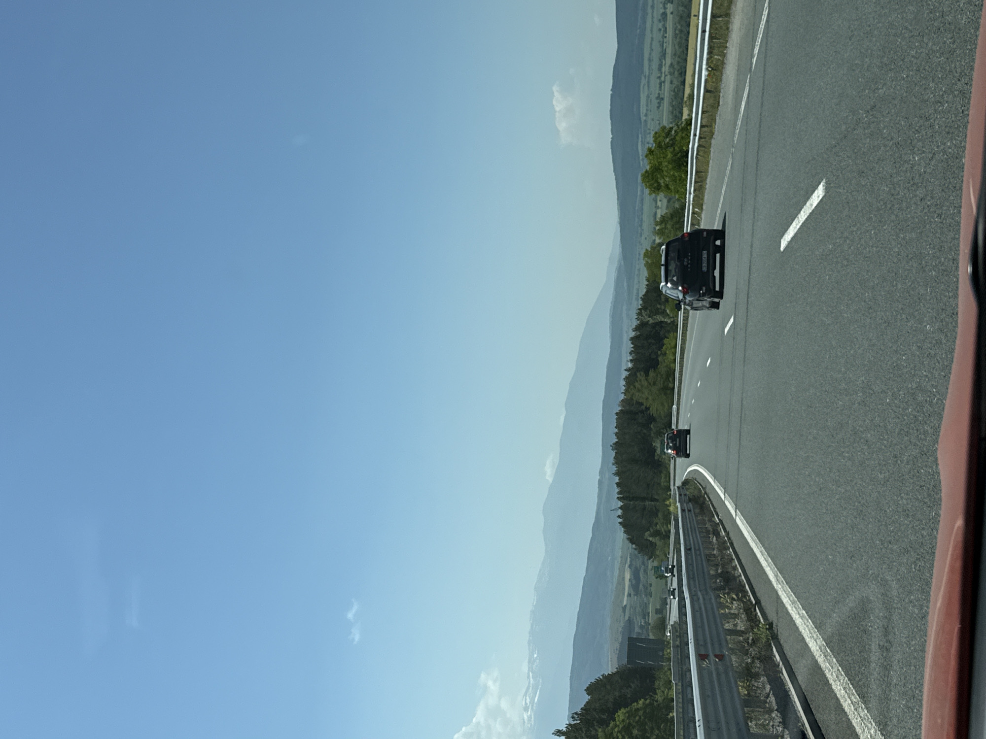 The image captures a sunny day on a multi-lane highway near Staro Selo, Bulgaria, viewed from the interior of a moving vehicle. The bright blue sky occupies the upper two-thirds of the frame, featuring a few faint, wispy white clouds. In the mid-distance, a range of mountains stretches across the horizon, with some distant peaks showing patches of snow or light rock and appearing faded due to atmospheric haze. Below the mountains, rolling hills covered in a mix of dense evergreen and deciduous forests are visible, transitioning into open, possibly cultivated, green fields in the valleys.
The asphalt highway features at least two lanes in the direction of travel, marked by white dashed lines, and a solid white line on the left shoulder. Metal guardrails line both sides of the road, separating it from the surrounding terrain. Several vehicles are present on the highway. In the immediate foreground, a black SUV (possibly a Hyundai) travels in the right lane, with its rear clearly visible. Further ahead in the left lane, a dark-colored sedan or smaller SUV is seen. Additional vehicles appear as dark specks in the distance along the highway. The bottom edge of the frame reveals a portion of a red dashboard or vehicle interior, indicating the viewer's position within a car. The overall scene suggests travel through a mountainous and rural landscape under clear weather conditions.