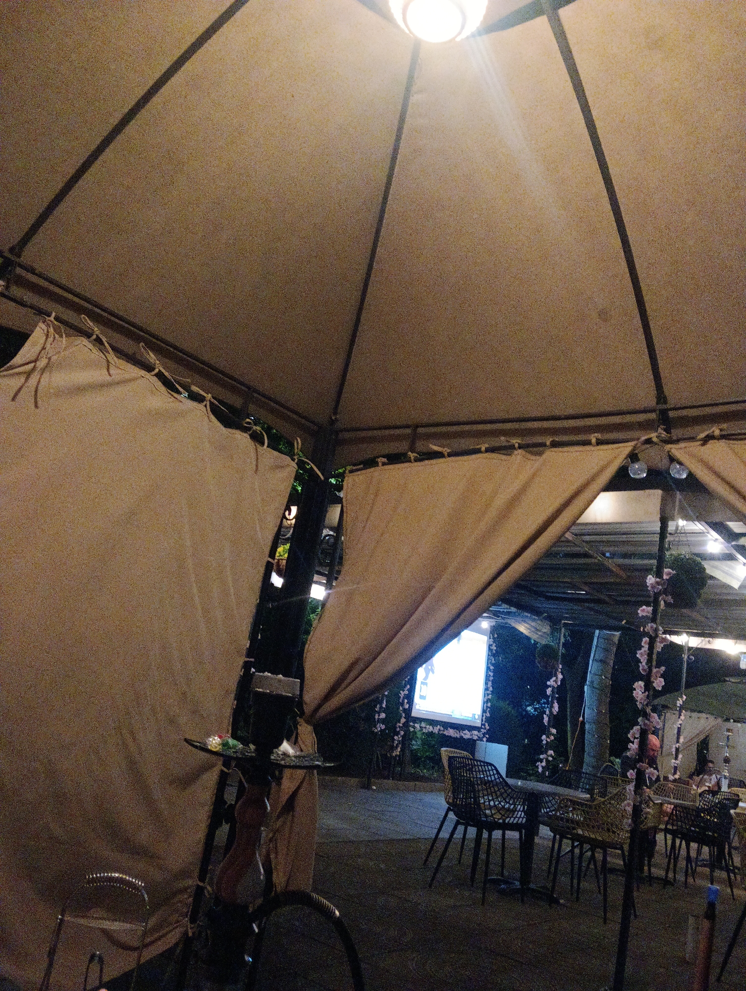 The image depicts an outdoor seating area, possibly a shisha lounge or a restaurant, under a large canopy. The canopy is made of a light-colored fabric stretched over a frame, supported by poles. Drapes of similar fabric are drawn to one side, revealing a view of tables and chairs arranged on a paved surface. In the background, a large screen is illuminated, and several people are seated at tables. Decorative elements, such as pink flowers on branches, are visible. A hookah is positioned in the foreground, near a chair. The overall ambiance suggests an evening setting, with artificial lighting illuminating the scene. The location is identified as Burgas, Bulgaria, though no specific landmarks or identifiers are visible in the image to confirm this.