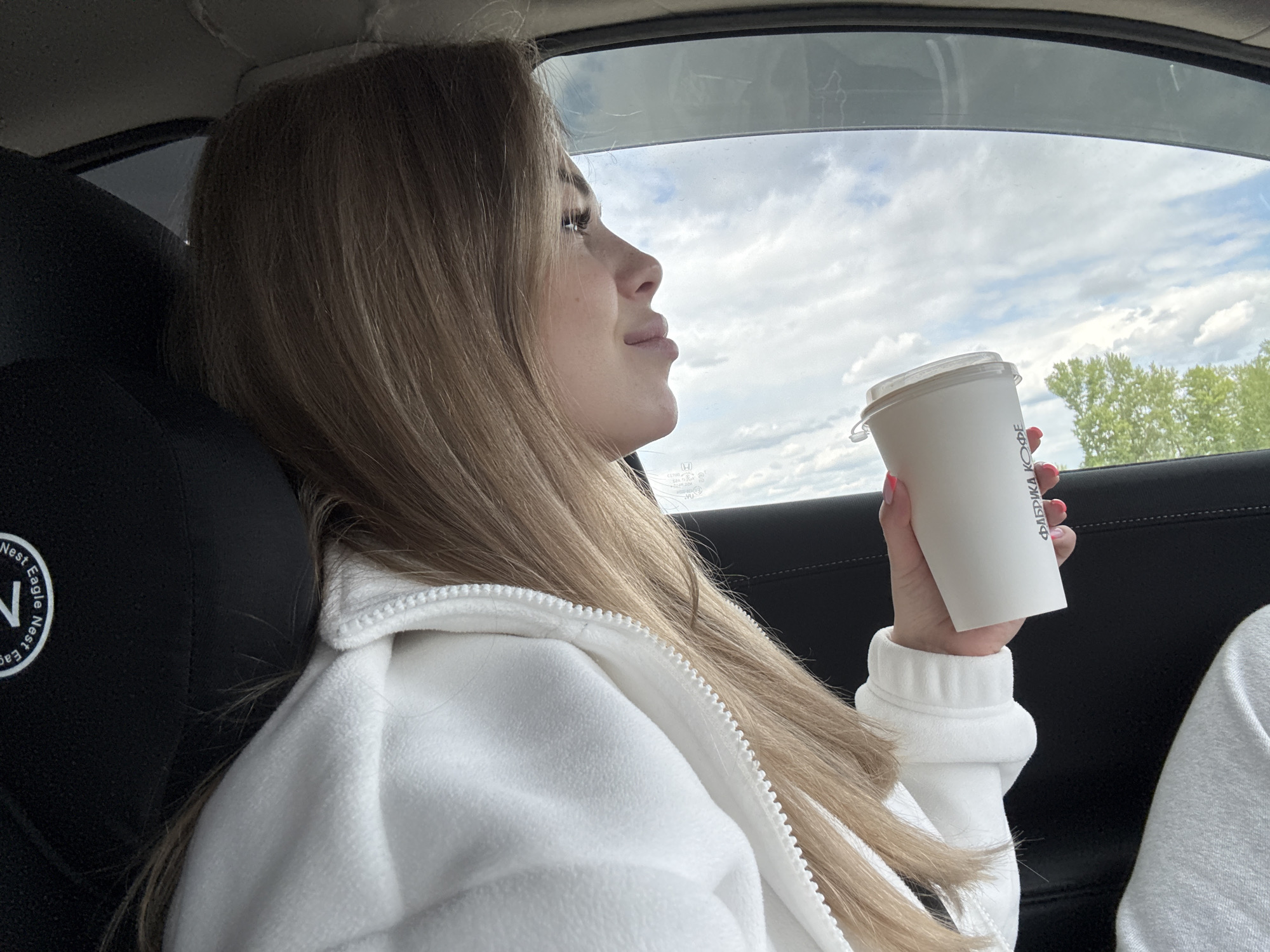 A young woman with long, light brown hair is sitting in a car, holding a white paper coffee cup in her right hand. She is wearing a white zip-up jacket with ribbed cuffs. The woman is looking out the car window, which shows a cloudy sky and trees in the distance. The car's interior is visible, including a black headrest with "Nest Eagle" printed on it and the car door panel. The coffee cup has text in a language other than English.
