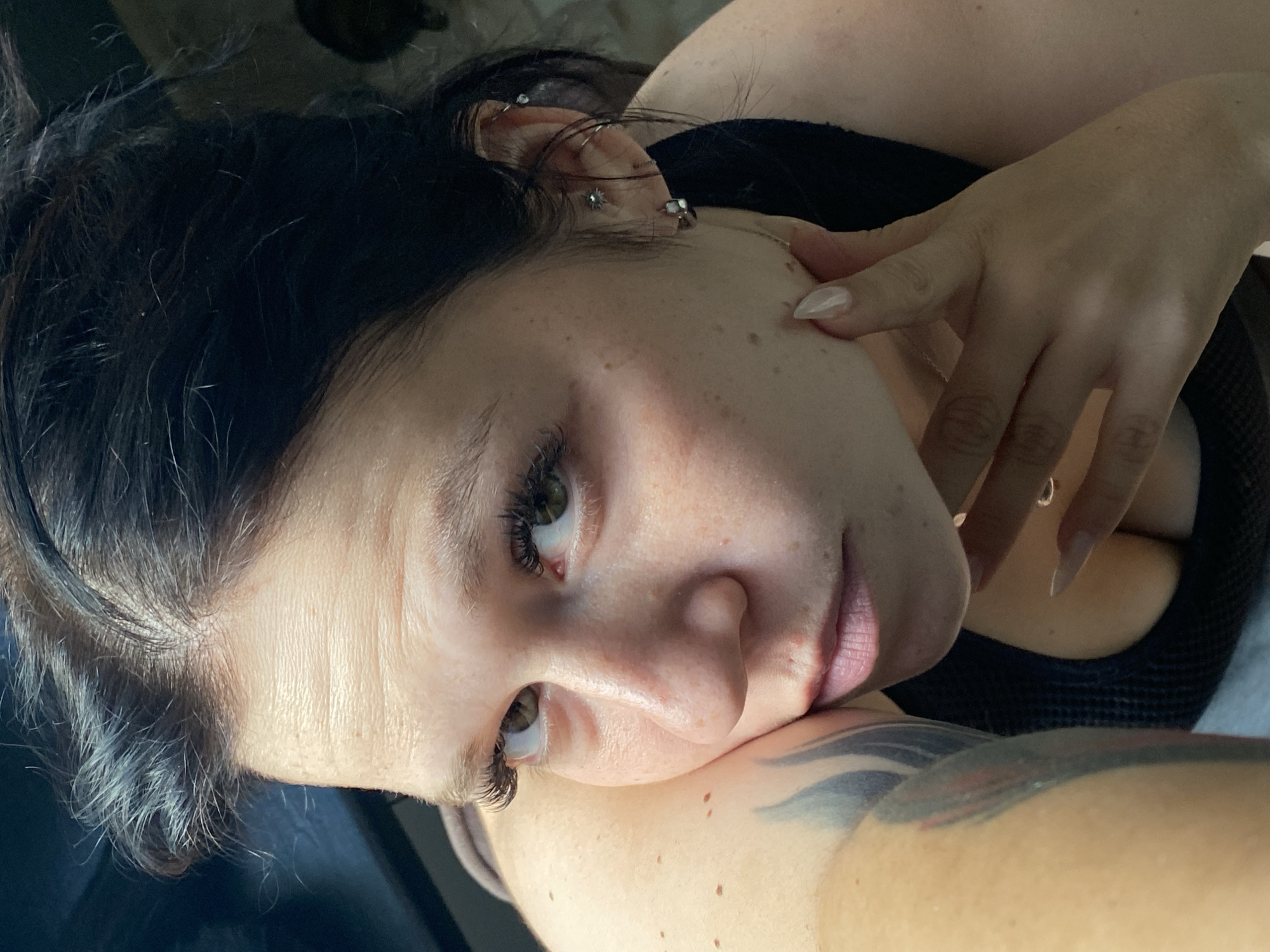 A close-up, high-angle, medium shot captures a woman from the shoulders up, her head tilted to the left. She has dark hair pulled back, revealing her face and left shoulder. Her eyes are green and gaze directly at the camera, framed by long, dark eyelashes. Her skin is fair with visible freckles and slight redness on her cheeks. She is wearing a dark-colored tank top, with her cleavage partially visible. Her left arm rests on her cheek, and her right hand, with long, light-colored fingernails, is positioned near her face, touching her chin. She has multiple earrings in her left ear, including hoops and studs. A tattoo is visible on her left upper arm, featuring abstract black and gray designs. The background is dark and out of focus, offering no discernible details about the environment or location. There is no information to indicate this image is located in Chaikovsky, Russia.