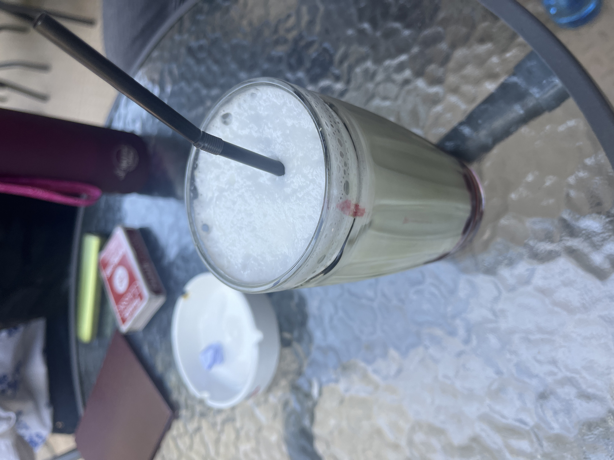 A close-up shot captures a glass of frothy, pale green liquid with a black straw inserted into it, resting on a textured glass table. A red mark is visible on the exterior of the glass near the rim. To the left, a small white ashtray holds a light purple object. Further left, a deck of playing cards with red detailing and a brown booklet or notebook are present. In the background, a dark purple bottle with a pink strap and a portion of a person's arm are visible, as is a hint of the outdoor environment with shadows and metallic chairs.