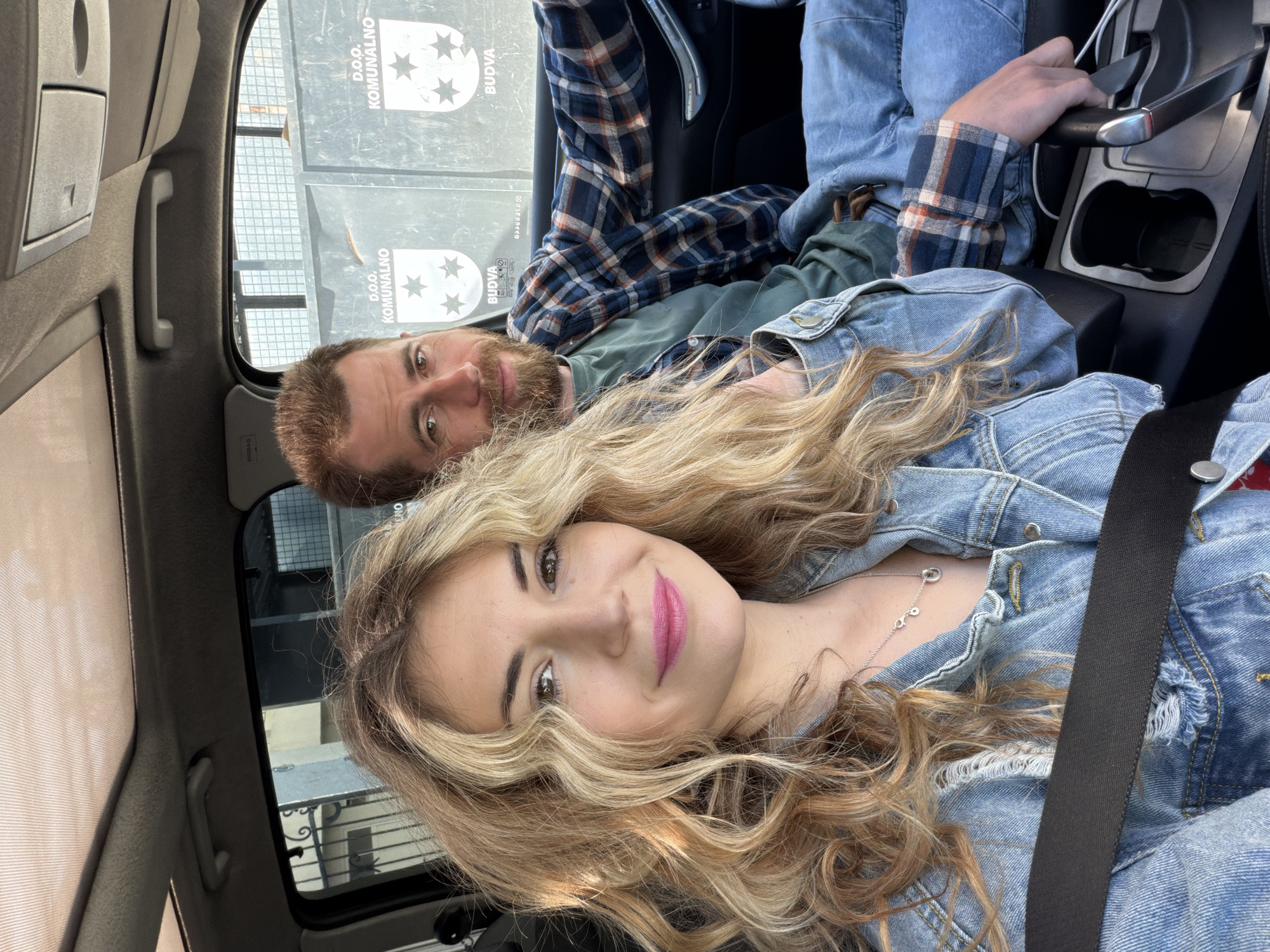 Inside a car, a woman with long blonde hair wearing a denim jacket and a seatbelt is in the foreground, smiling towards the camera. A man with a beard, wearing a plaid shirt, is in the background. Through the car window, a building with "D.O.O. KOMUNALNO BUDVA" is visible, suggesting a location in Budva, Montenegro. The man in the background has his hand near the car's gear shift.
