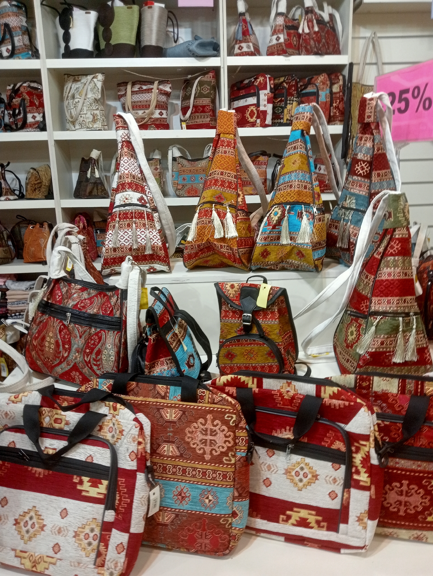 This is a photograph of a retail display featuring a variety of bags with colorful, ethnic-inspired patterns, likely taken in Korkuteli, Turkey. The bags are arranged on white shelves and a white countertop. The items include tote bags, backpacks, and shoulder bags, all adorned with intricate geometric designs and tassels. The background shows more shelves with additional bags, and a bright pink sign with "25%" printed on it is visible on the right side of the image. The lighting is bright and even, suggesting an indoor retail environment. There are no people visible in the photograph.