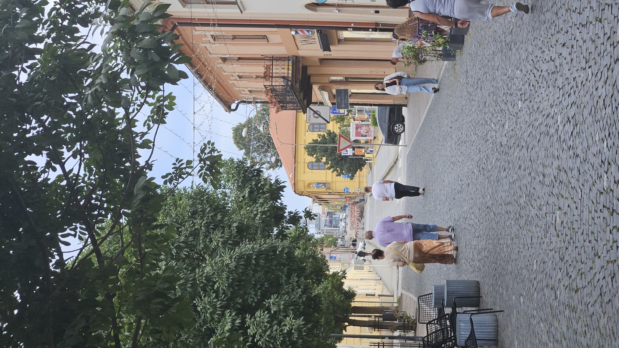 The image shows a street scene in Prešov, Slovakia, likely in the historic city center, with cobblestone paving and buildings lining both sides of the street. In the foreground, three people are walking away from the camera on the sidewalk. The person on the left is wearing a light-colored top and a long, flowing brown skirt, carrying a light-colored bag over their shoulder. The person in the middle is wearing a light purple short-sleeved shirt and blue shorts. The person on the right is wearing a white t-shirt and black pants.
Further down the street, on the right side, several shops are visible. One shop has a barber pole outside and the sign "VAM" above its entrance. Another building has a balcony with flowers. On the left side of the street, there are trees with dense green foliage casting shadows on the sidewalk. Some outdoor seating with tables and chairs is visible on the left sidewalk.
In the background, more buildings, including a yellow building with blue statues on its exterior, are visible. There are also vehicles, traffic signs, and streetlights. The sky is overcast with some light visible. The overall impression is a somewhat busy, pedestrian-friendly street in a European city.