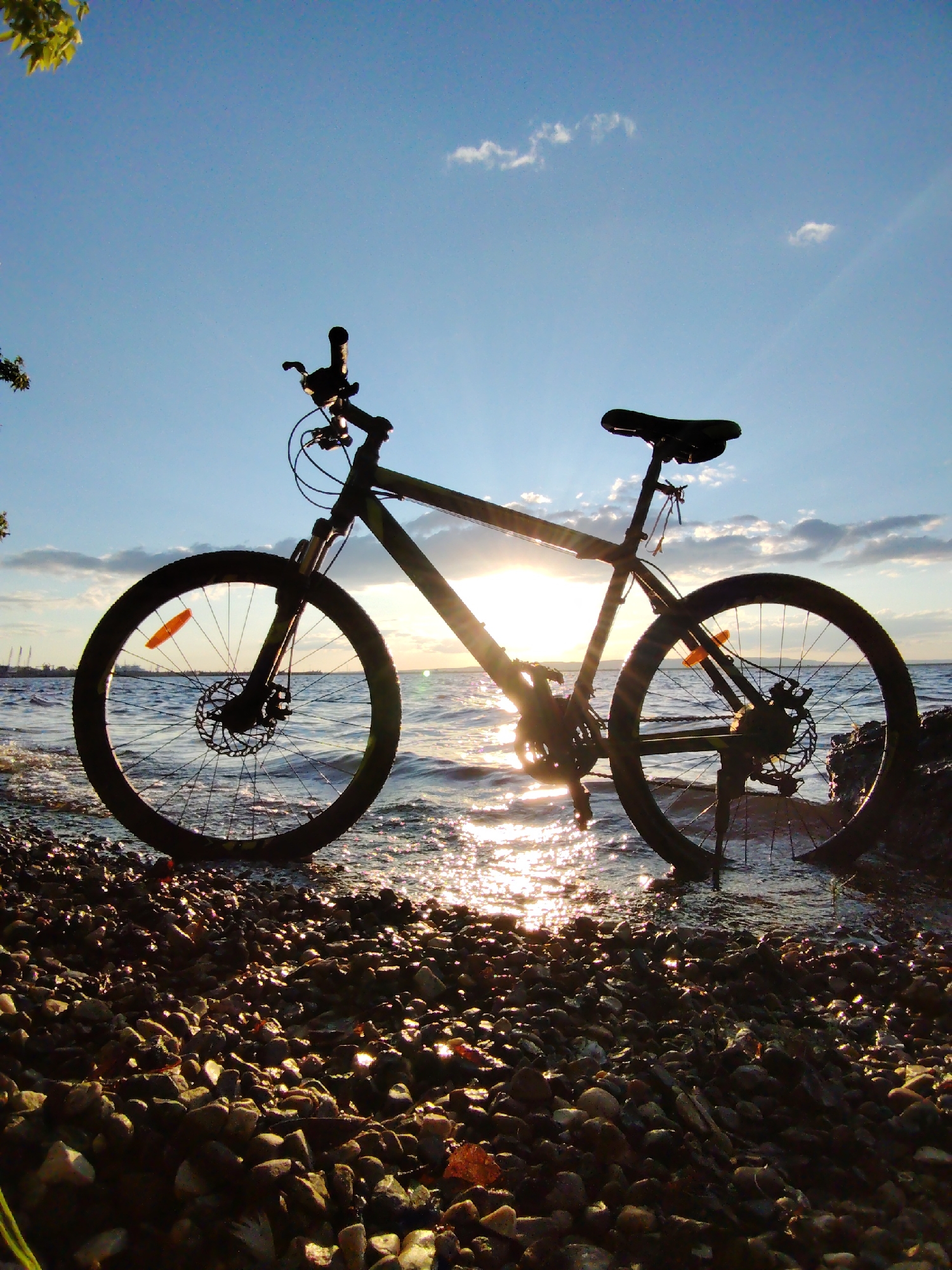 A black bicycle is silhouetted against a setting sun, positioned on a rocky shoreline. The bicycle's front wheel is partially submerged in the water, and its tires are resting on a mix of small and large stones. The sun is low on the horizon, casting a warm glow and creating a shimmering reflection on the water's surface. The sky is a clear blue with a few wispy clouds. The background shows a body of water with industrial structures visible in the distance. The scene is captured from a low angle, emphasizing the bicycle and its interaction with the natural elements. The location is identified as the Kama River embankment in Chaikovsky, Russia.