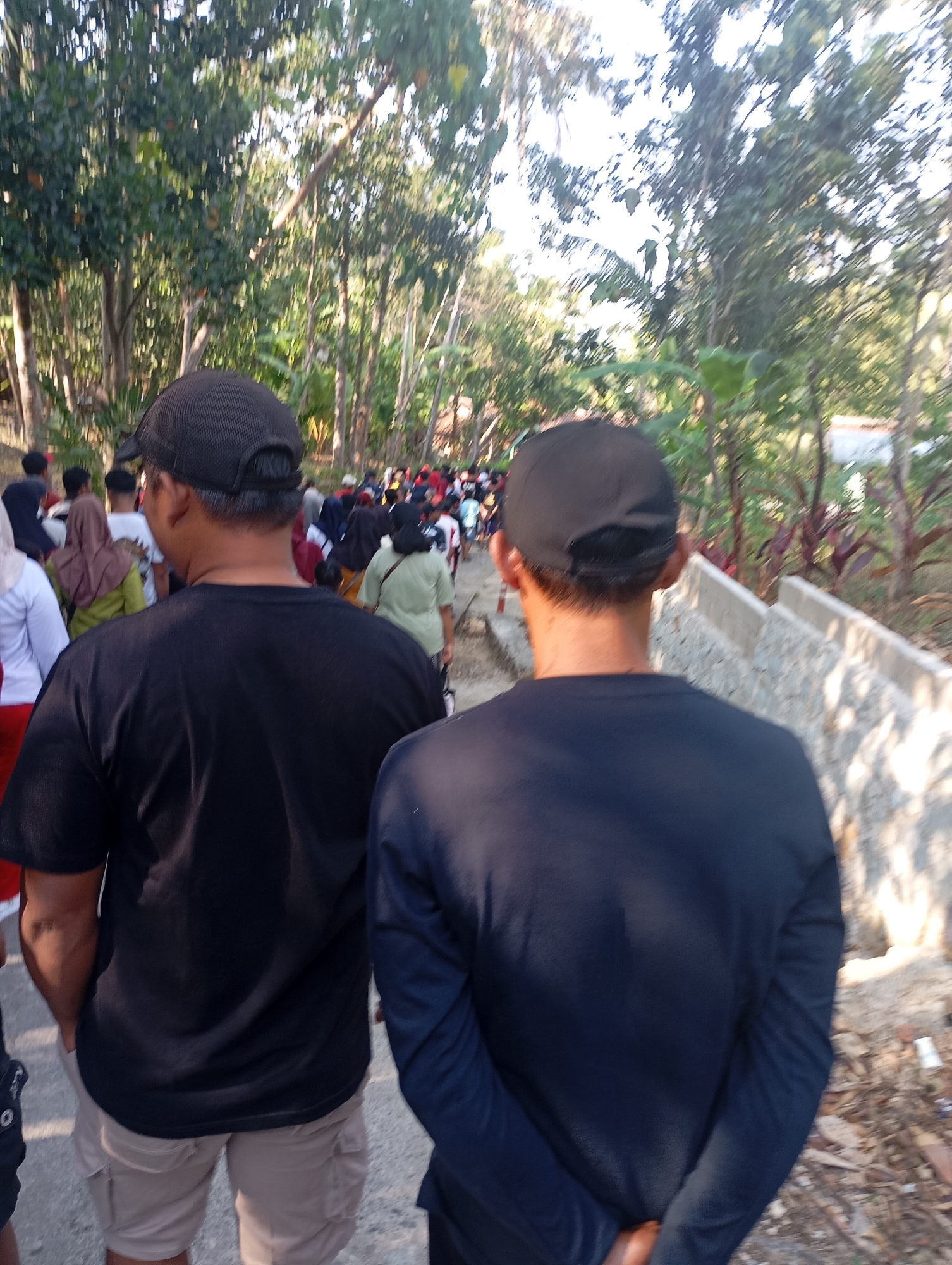 In Ketepung, Indonesia, a group of people walk along a dirt path lined with trees and foliage. Two men, viewed from behind, are in the foreground. The man on the left wears a black t-shirt and tan cargo shorts, while the man on the right wears a navy long-sleeved shirt. Both men wear black caps. The path continues into the distance, where more individuals are visible, suggesting a procession or gathering. The environment appears to be rural or semi-rural, with a rustic wall constructed from stone and concrete along the right side of the path. Sunlight filters through the trees, casting shadows.