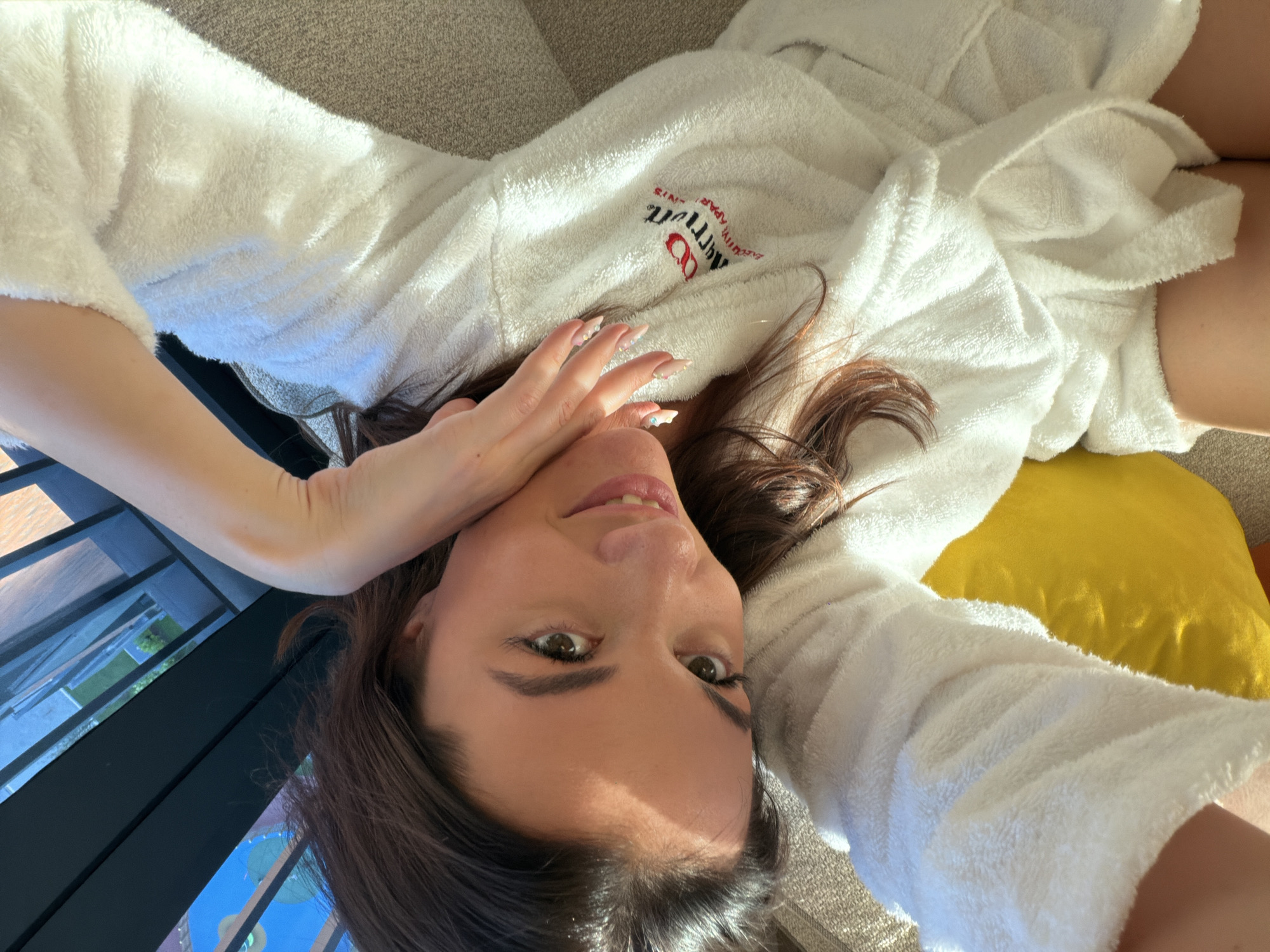 A woman with light skin and brown hair is lying on her back, wearing a white robe with an "Arrivato Apartments" logo on the left chest. Her left hand is raised to her face. She is lying on a gray couch with a yellow pillow. A black balcony railing is visible in the background. The location is within or close to Küçükçekmece, Turkey, based on the branding of the robe.