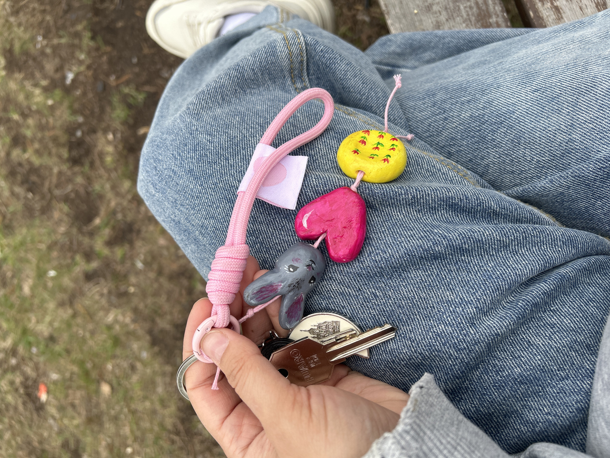 A person sits on a wooden bench in Uray, Russia. The individual wears blue jeans and white sneakers. In their hand, they hold a keychain with several decorative charms, including a yellow circle with painted red flowers, a pink heart, a gray elephant head, and various keys, including a "GUARD" key. The setting appears to be outdoors, with a blurred ground area visible in the background.