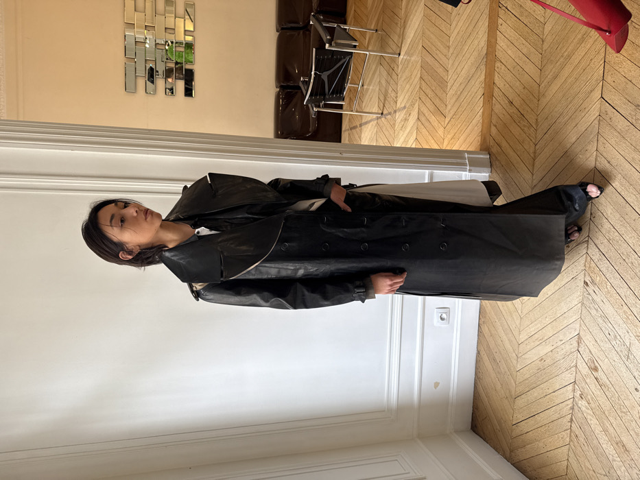 The image shows an interior scene within what appears to be a Haussmannian-style apartment or building, characteristic of Paris, France. An adult individual with short, dark hair, wearing a long black leather trench coat over a lighter garment and black open-toed sandals, stands slightly left of center in the foreground. Their right hand is in the coat pocket, and their gaze is directed towards the right.
The immediate room features white walls with decorative molding (wainscoting) and light-toned herringbone parquet flooring. An electrical outlet is visible on the wall. Through an open doorway to the right, a second room is visible. This room has light beige walls, a geometric mirror art installation composed of rectangular segments, and dark leather seating, including a modern chair with a chrome frame and black leather elements. A reflection in the mirrors shows a person in a bright green top. A small portion of a red bag is visible in the bottom right corner of the frame.
