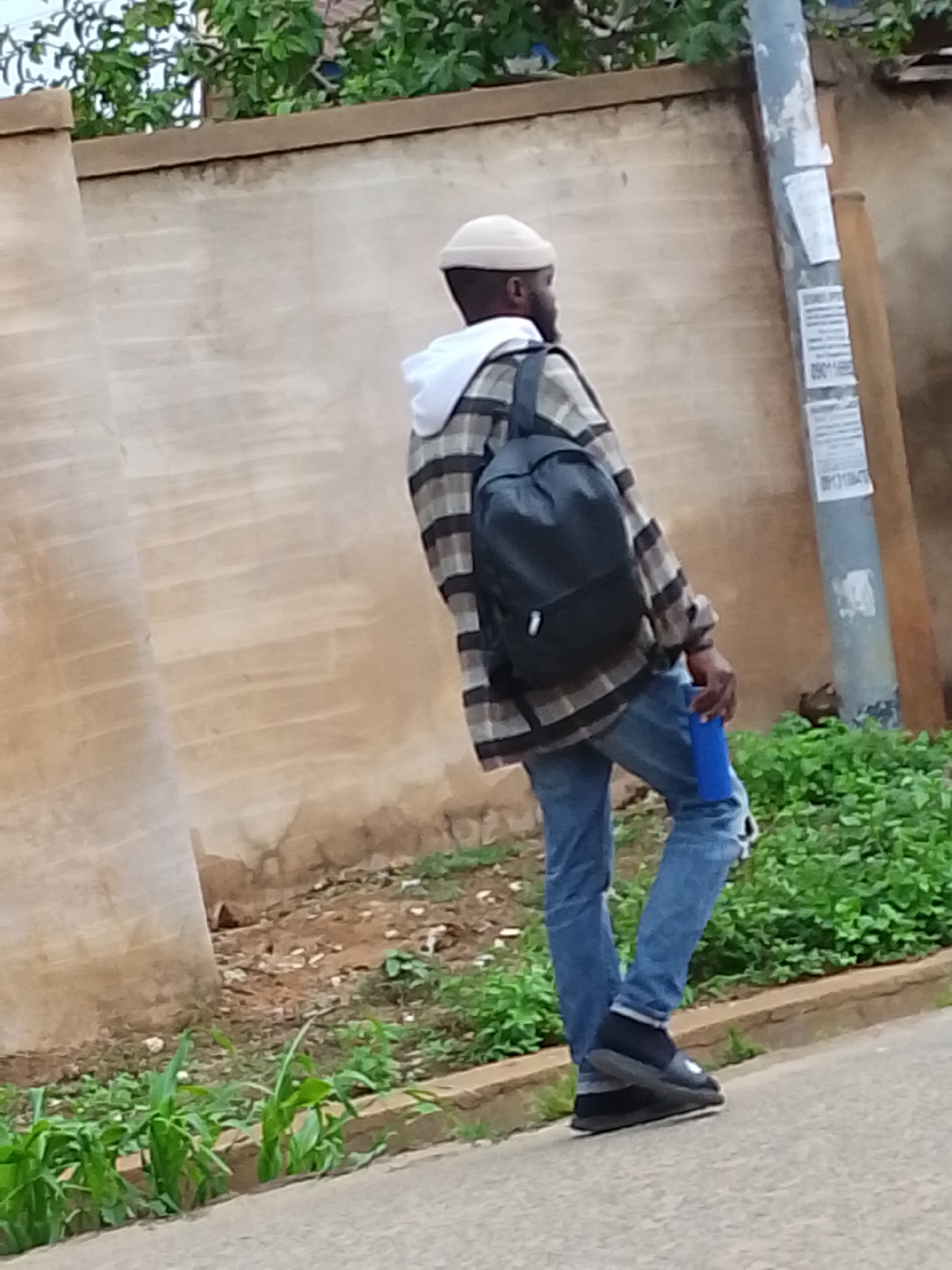 A person stands outdoors, facing to the right. They wear a light-colored beanie, a white hoodie, a plaid shirt, blue jeans with a tear in the knee, and black slip-on shoes with socks. A black backpack hangs on their back, and they hold a blue object in their right hand. They stand on a paved surface near green vegetation and a concrete curb. A wall and a utility pole are in the background. The location is in a city of Unknown, Unknown.