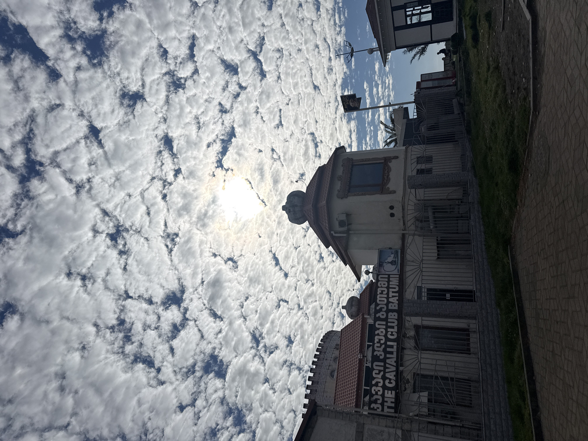 The image displays an outdoor, daytime scene in Batumi, Georgia, with a strong focus on the sky and adjacent buildings. The sky is a prominent feature, blanketed by a dense pattern of numerous small, white, fluffy clouds (likely altocumulus or cumulus humilis) against a backdrop of blue sky. The sun is visible in the upper left quadrant, partially obscured by the clouds, indicating a bright day.
On the right side of the frame, several buildings are visible. One distinct building features a reddish-brown tiled roof and an ornate, crown-shaped decoration atop its peak. An air conditioning unit is mounted on its facade. Adjacent to this, another building prominently displays a large sign that reads "THE CAVALLI CLUB BATUMI" in English, alongside Georgian script.
The ground level shows a paved area, likely a sidewalk or street, with a strip of green grass running along the edge of the properties. Fencing and gates are also visible in front of some structures. A street lamp with an unusual antenna-like fixture is positioned between the buildings. The scene appears devoid of people or specific activities, creating a sense of quiet observation. The bright sunlight filtering through the clouds suggests it is midday or early afternoon.