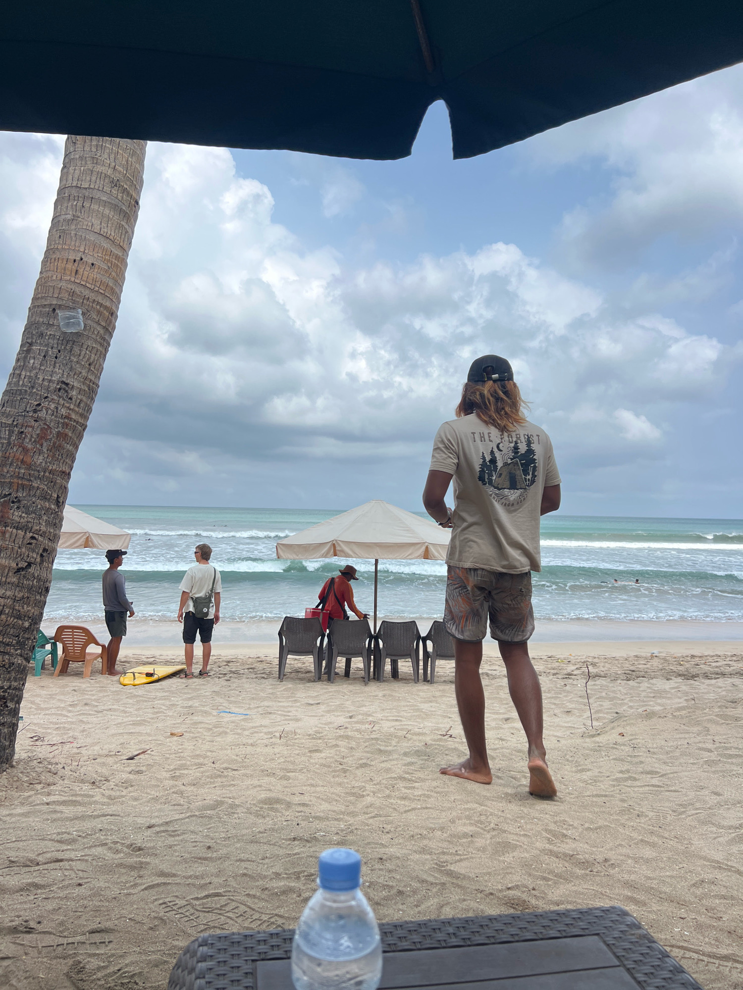In Kuta, Indonesia, a beach scene unfolds under a cloudy sky. In the foreground, a person with long blonde hair and a cap stands facing away from the camera, wearing a tan t-shirt with a graphic of a cabin and trees, and patterned shorts. A bottle of water sits on a dark, textured table in front of them.
Further back on the sandy beach, two men stand talking near a yellow surfboard. One man wears a grey long-sleeved shirt and dark shorts, the other a light-colored t-shirt and dark shorts. A third person, wearing a red shirt and hat, is near a cluster of dark chairs and two beige umbrellas. The ocean with moderate waves is visible behind them, with a few surfers in the distance. A prominent palm tree trunk stands on the left side of the frame, with a dark umbrella overhead. The overall atmosphere is casual and relaxed, indicative of a beach day.