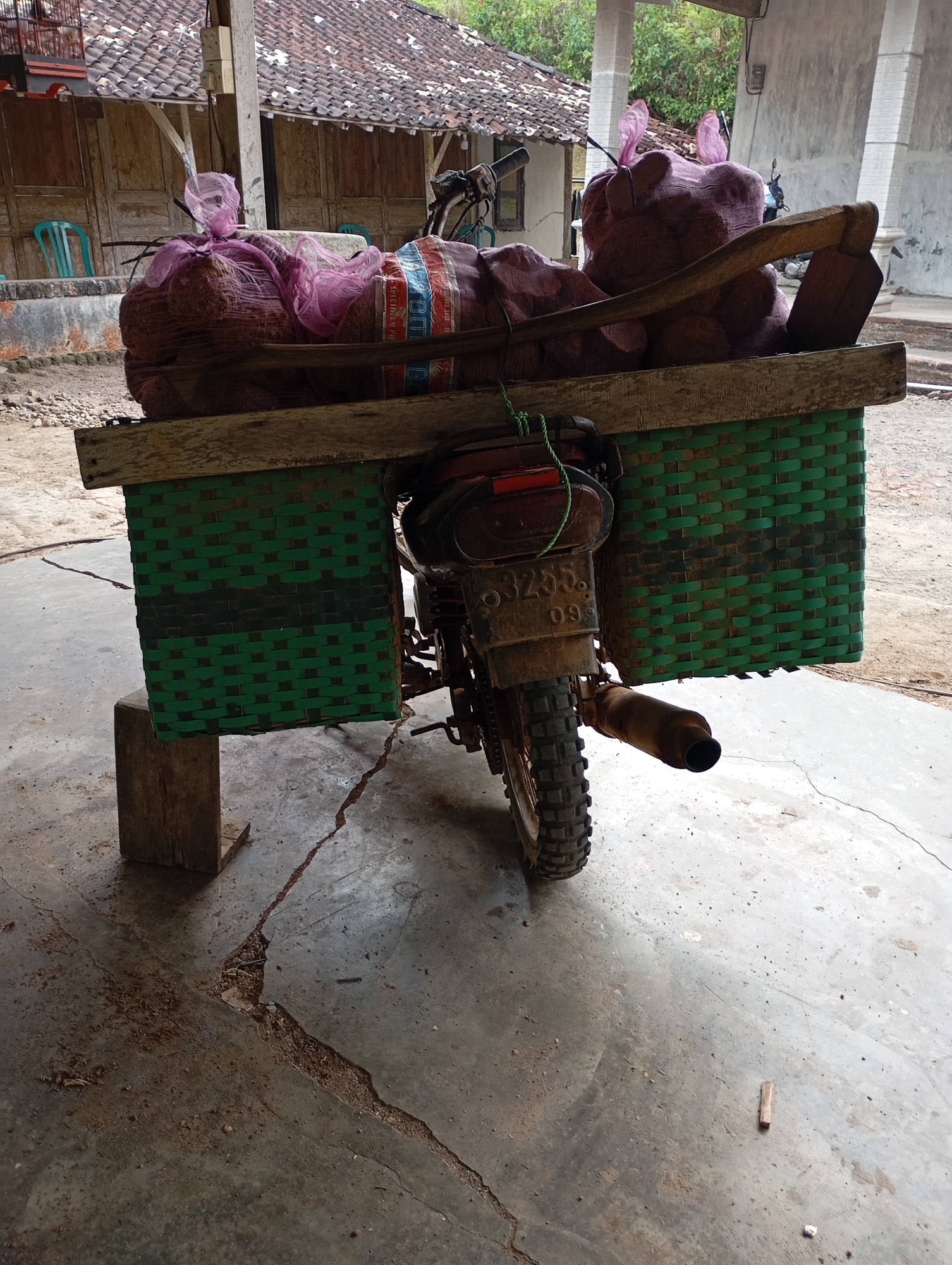 The image depicts a close-up view of a modified motorcycle in Ketepung, Indonesia. The motorcycle has a large, woven green basket attached to its rear, serving as a cargo carrier. This basket is laden with several large, burlap sacks filled with a reddish-brown substance, possibly produce like onions or potatoes, secured with pink netting and ropes. A wooden plank rests on top of the sacks, extending across the basket. A wooden handle of a tool, possibly an agricultural implement, is visible protruding from the sacks. The motorcycle itself is a dirt bike model with knobby tires and a visible exhaust pipe. The rear license plate reads "3255 09". The scene is set on a concrete ground with visible cracks and debris. In the background, a traditional Indonesian house with a tiled roof and wooden walls is visible, along with some green plastic chairs and trees. The overall impression is one of rural utility and transportation.