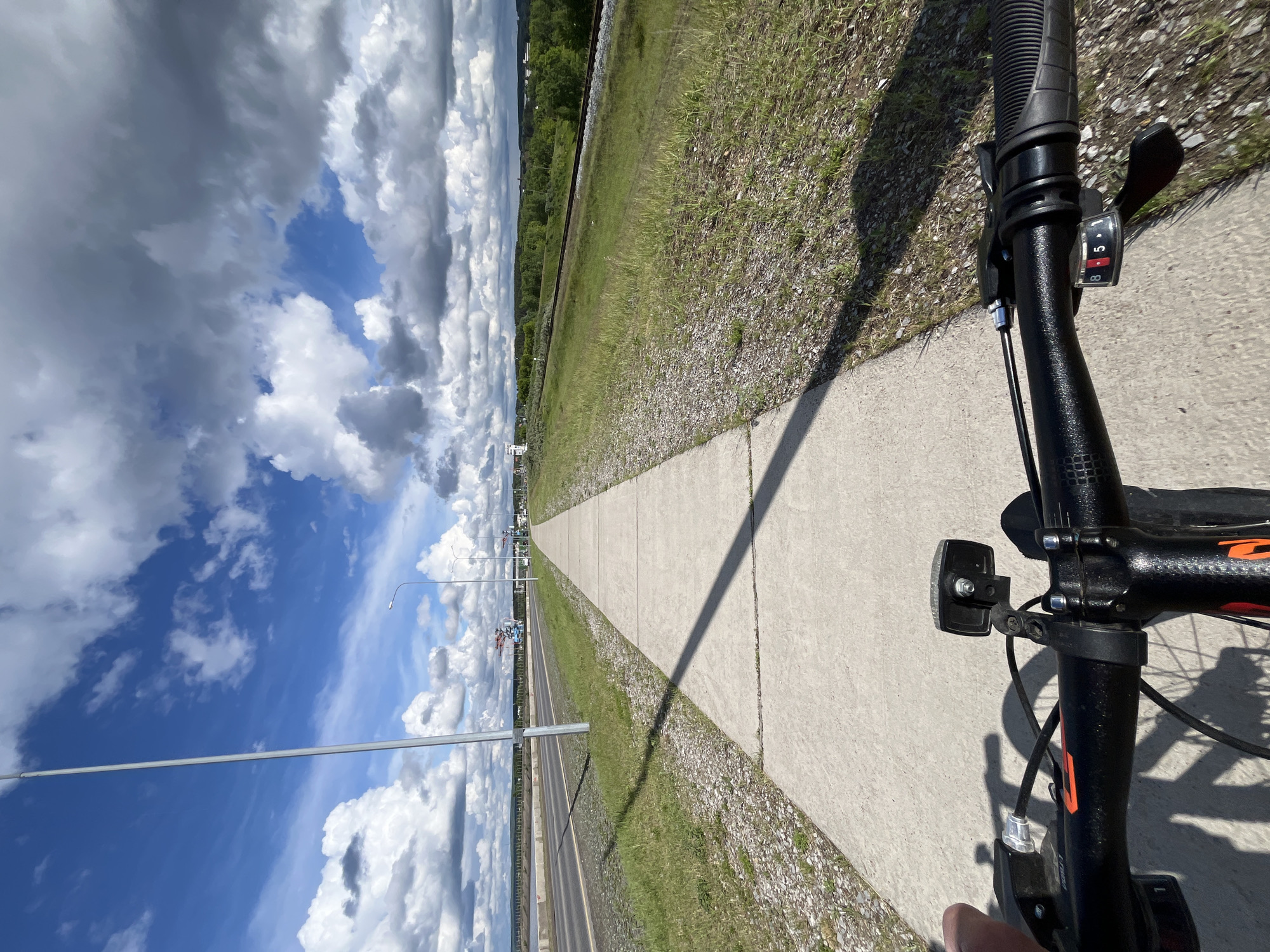 The image captures a first-person perspective from a bicycle, looking forward along a concrete path. In the foreground, the black handlebars of a bicycle are visible, featuring a textured rubber grip on the right, a black gear shifter displaying the numbers "8" and a highlighted "5" (indicating gear selection), and a small black front light mounted to the stem. Part of a hand or finger is visible at the bottom right.
The concrete path extends into the distance, bordered on both sides by grassy verges with patches of gravel. To the left of the path is a multi-lane road or highway, also stretching into the distance and lined with streetlights. Beyond the road, in the mid-ground, there's a more extensive grassy area, and further back, a train track or similar railway line runs parallel to the road. Rolling hills covered in green trees are visible on the horizon.
The sky is a prominent feature, filled with large, fluffy white and grey cumulus clouds against a vibrant blue background. Strong, elongated shadows from the bicycle and possibly from an unseen structure stretch across the path from the right, suggesting bright daylight, possibly morning or late afternoon. The overall scene depicts a sunny day in an open, semi-urban or rural environment in Chaikovsky, Russia, during a bicycle ride.