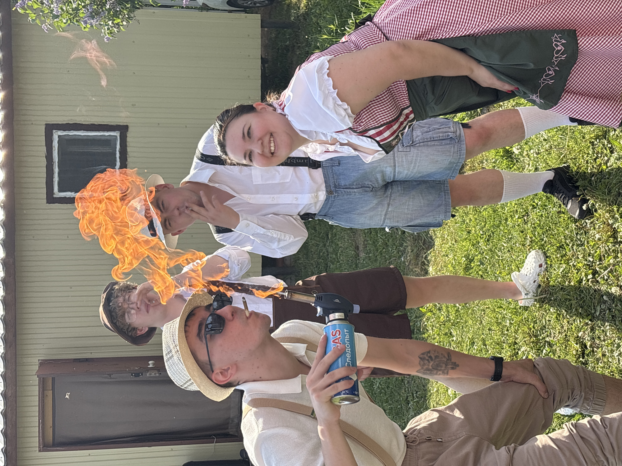 The photograph shows four people outdoors. One person in the foreground is holding a blowtorch to a cigarette in their mouth, creating a large flame. The person is wearing a light-colored outfit, a straw hat, and sunglasses. Behind them, another person wearing a similar outfit and hat has a small flame near their mouth. A third person is further in the background. A fourth person, wearing a red and white dress and white stockings, smiles at the camera. They are all outside on a grassy area, with a building behind them and a vehicle visible in the background. The blowtorch canister has lettering in a Cyrillic script, including the word "следопыт". I have no basis to determine if this photo was taken in Пальники, Russia.