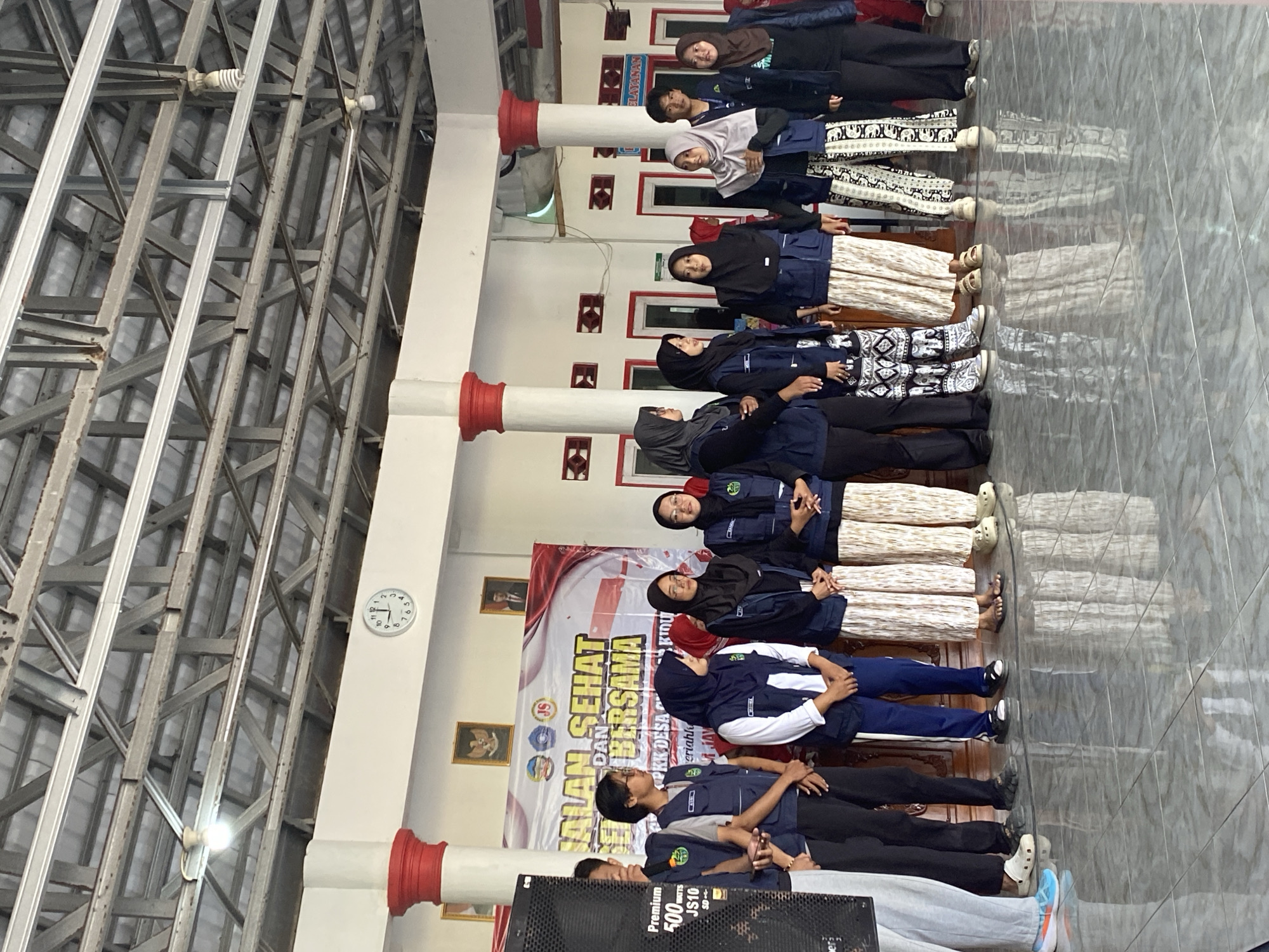 In the city of Gumelar Kidul, Indonesia, a group of approximately 13 individuals, primarily young adults, stand in a line on a tiled floor. They are positioned in front of a white banner with red and black text that reads "JALAN SEHAT DAN BERSAMA". Many of the individuals are wearing dark blue vests over their clothing, and several are wearing hijabs. Some are looking forward, while others are looking to the side. The environment appears to be an indoor space with a high ceiling featuring exposed metal beams and lighting fixtures. On the wall behind them, a circular clock shows the time as approximately 10:13. There are also framed pictures on the wall. To the left of the group, a large black speaker is visible. The scene suggests an event or gathering taking place.