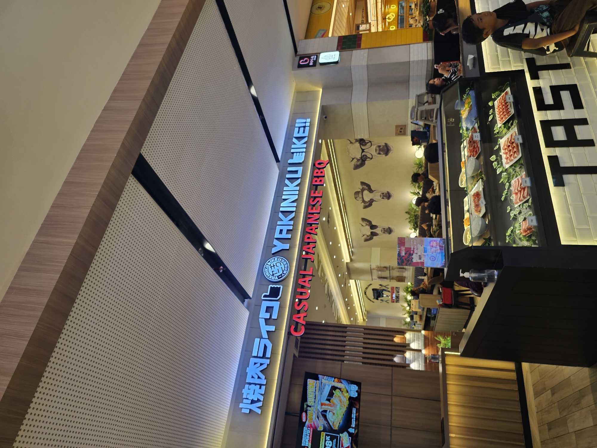 The image shows the exterior of a Japanese BBQ restaurant called "YAKINIKU LIKE!!" in Condongcatur, Indonesia. The restaurant features a prominent blue neon sign with Japanese characters and English text. Below the main sign, a smaller red sign reads "CASUAL JAPANESE BBQ." The restaurant's interior is visible through large glass windows, revealing a display case filled with raw meat and vegetables, suggesting a self-service or customizable dining experience. Several customers are seated at tables inside, and some staff members are visible behind the counter. A young boy is seated on a stool in the foreground, facing away from the camera. The overall ambiance appears to be modern and clean, with warm lighting.
