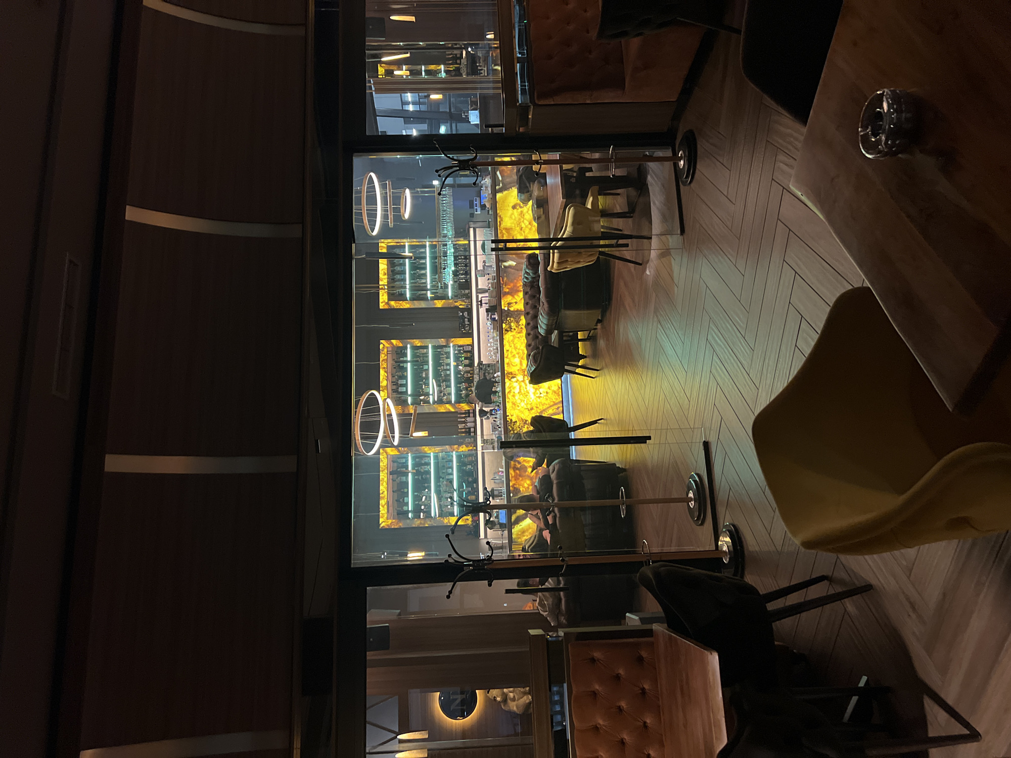 This image depicts the interior of a restaurant or bar, possibly in Burgas, Bulgaria, as suggested by the prompt. The viewpoint is from a seating area with a wooden table and chairs (one yellow), looking out through a glass partition. Beyond the glass is a bar area, with shelves of liquor bottles illuminated with yellow lighting, suggesting an upscale or modern aesthetic. There are ring-shaped lights hanging near the partition. A coat rack stands near the glass. The room's overall lighting is dim. Several people are visible through the partition, seated at tables or near the bar. The floor is a light-colored wood arranged in a herringbone pattern. A dark ash tray is visible on the table. The seating area appears relatively empty, except for the table in the foreground. There are brown upholstered chairs, and possibly other tables and chairs.