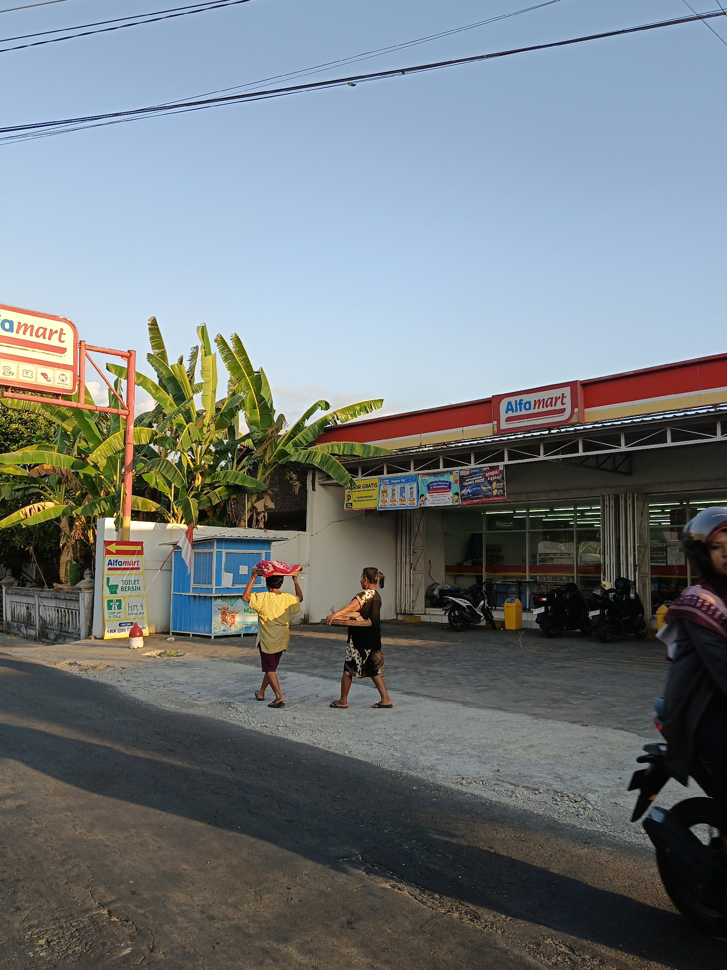 This image depicts an outdoor scene in front of an Alfamart store, a convenience store chain, in Trucuk, Indonesia. The sky is clear blue, with power lines running horizontally across the upper portion of the frame. Banana trees with large green leaves are visible behind the store's signage.
In the foreground, a paved road is in the lower half of the image, with a distinct shadow cast across it, indicating the time of day is likely late afternoon or early morning. To the right of the road, a gray concrete pathway leads to the Alfamart store.
Two women are walking on the pathway. The woman on the left is wearing a yellow shirt and dark shorts, and she is carrying a tray of items on her head, covered with a colorful fabric. The woman on the right is wearing a dark patterned skirt and a dark top. She appears to be walking behind the first woman, holding a flat tray or board.
In front of the Alfamart store, there are several motorcycles parked. The store itself has a red and yellow facade with the "Alfamart" logo prominently displayed. Various advertisements are visible on the store's exterior walls. There is also a small blue kiosk with a red and white Indonesian flag on it, positioned to the left of the store entrance. A red and white circular object, possibly a traffic cone or marker, is situated near the kiosk. Another person, partially visible, is riding a motorcycle on the far right of the frame, wearing a helmet and a patterned scarf.