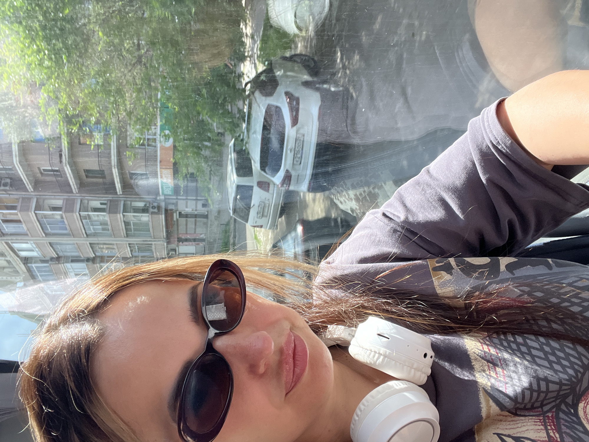In this close-up selfie, a woman with long, brown hair and sunglasses is shown from the chest up. She is wearing a grey t-shirt with a colorful graphic print and white headphones around her neck. The woman is looking towards the camera with a slight smile. The background is blurred, showing a building with multiple windows and balconies, green trees, and two white cars parked on a street. The scene appears to be outdoors on a sunny day, possibly taken from inside a vehicle due to the reflections on the glass.