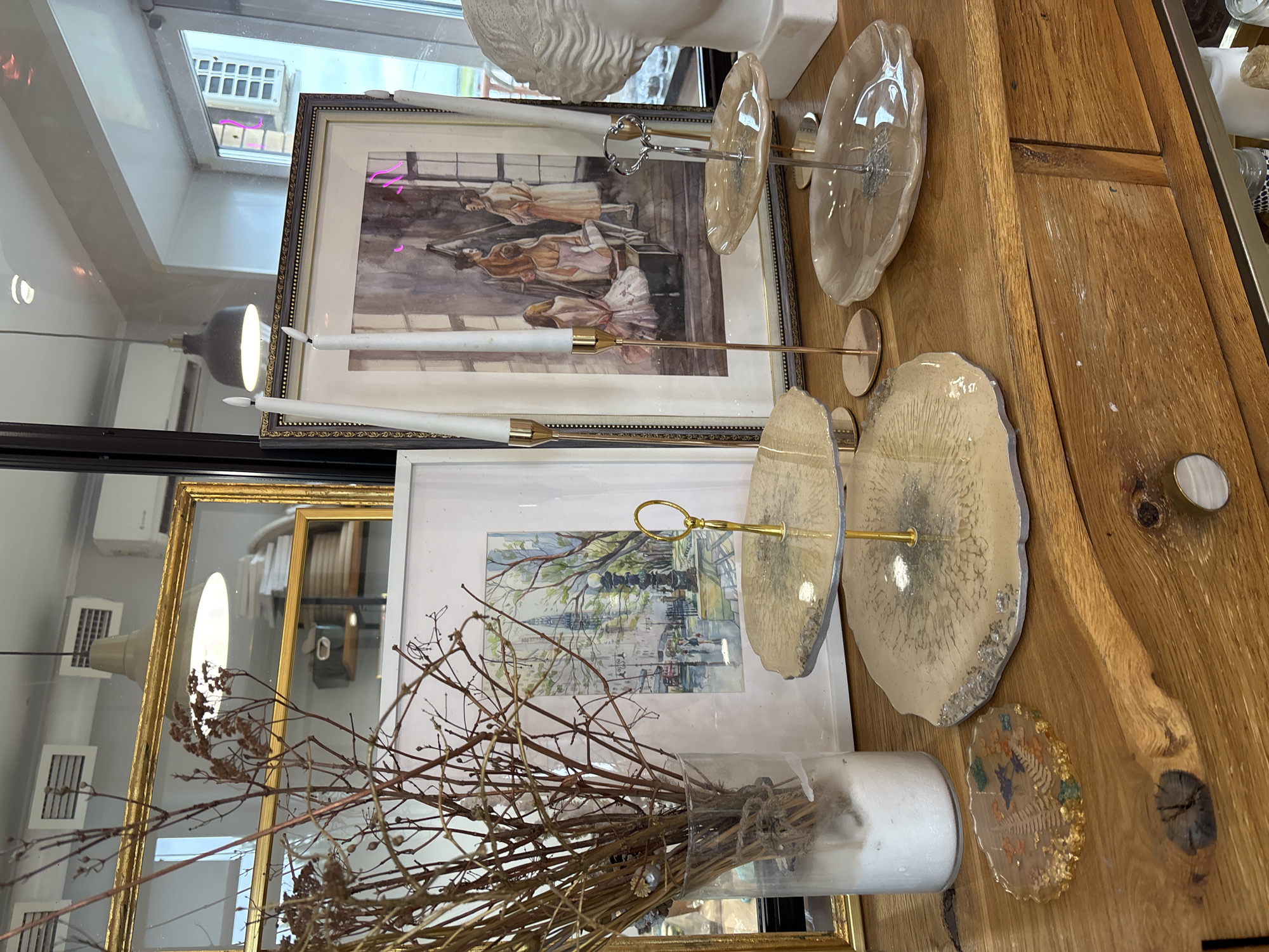 The image shows a wooden dresser with several decorative items. On the dresser are a glass vase filled with dried branches, a framed watercolor painting of a city street scene, two white taper candles in gold candlesticks, a two-tiered decorative serving stand with translucent, irregular-shaped plates, and a small, round decorative coaster with embedded dried flowers and glitter. To the right of the serving stand is a white bust of a classical figure. In the background, a large gold-framed mirror reflects lights and air vents on the ceiling, as well as a window with a view of a building. The overall scene appears to be an interior decoration display. The location is not discernible from the image, and there is no indication of it being in Naberezhnye Chelny, Russia.