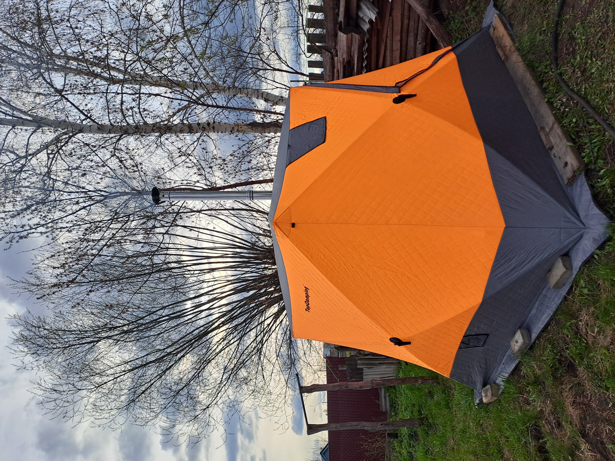 An outdoor scene captured in Polianki, Russia, features a large, hexagonal tent designed for cold weather, prominently positioned in the center. The tent is bright orange on its main panels and dark grey along its lower skirt and trim. A metallic chimney pipe extends vertically from the top of the tent, indicating it's equipped with a stove for heating, common for winter camping or ice fishing. The tent is secured to the ground, with concrete blocks and wooden planks visibly holding down its dark grey base skirt, suggesting a need for stability against wind or to keep warmth inside.
In the background, several deciduous trees with bare branches dominate the upper half of the image, their intricate networks of twigs silhouetted against a cloudy, overcast sky. The trees appear to be birches, identified by their white bark. The ground around the tent is covered with green grass, indicating spring or late autumn. A rustic wooden fence or structure is visible behind the tent, along with what appears to be part of a building in the distance. The lighting suggests it is daytime, possibly late afternoon or early morning, under a heavily clouded sky. The brand name "TopCamping" is visible in black text on one of the orange panels of the tent. No people are present in the scene. The overall impression is one of practical preparation for outdoor activities in a cool or cold climate.
