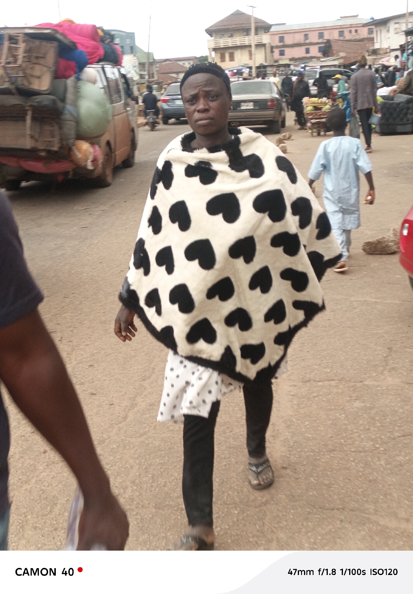 A medium shot captures a young woman in the foreground walking towards the camera. She wears a black and white patterned shawl with heart shapes and a black headband. She is also wearing a white dress with black polka dots underneath the shawl and black leggings. Her left arm is extended forward, holding what appears to be a plastic bag. In the background, a brown van is loaded with goods, and other people and vehicles are visible on a paved street, suggesting an urban environment, likely in Jos, Nigeria. The lighting suggests it is daytime.