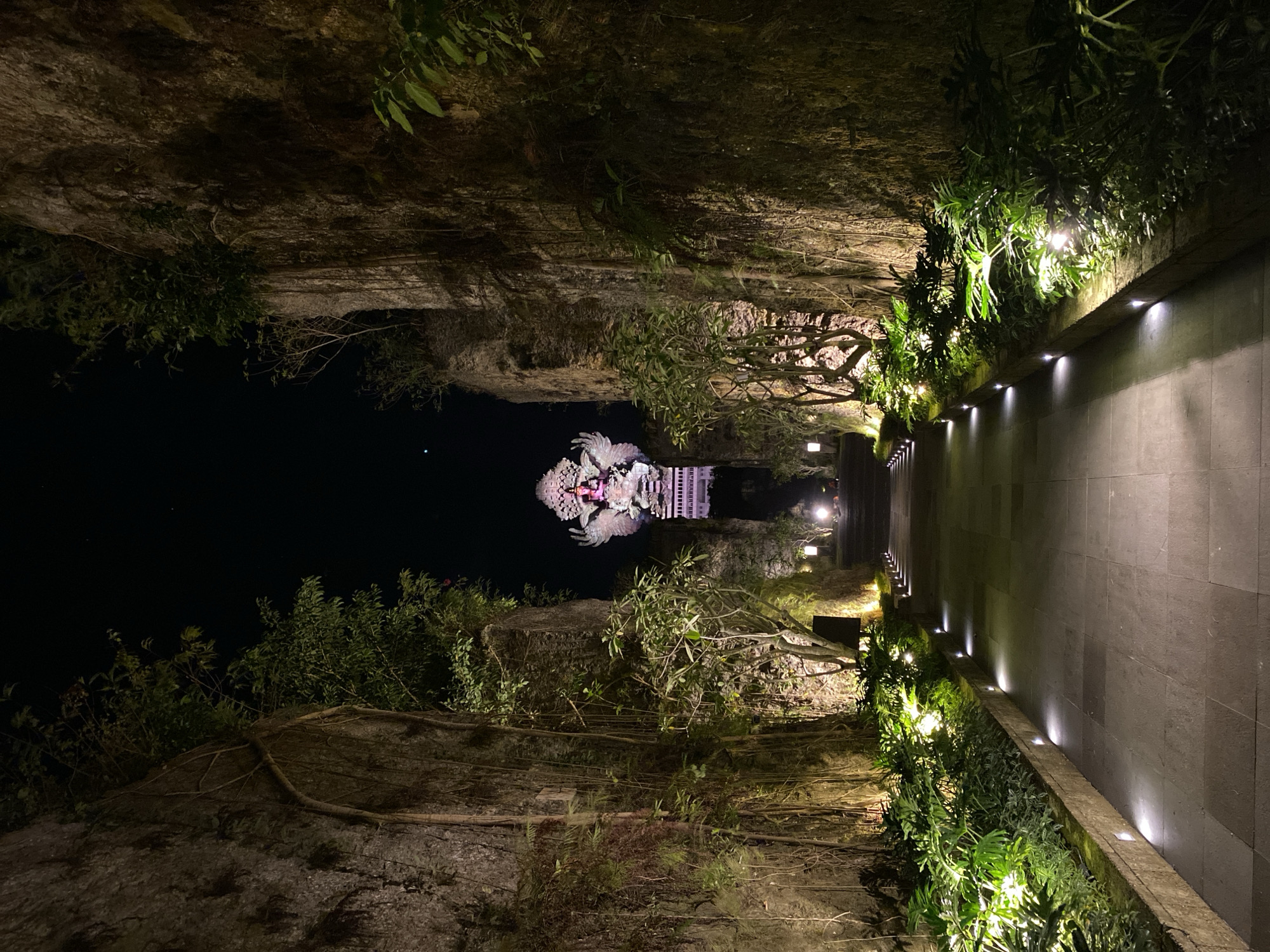 The image shows a pathway leading towards a large, illuminated statue at night. The pathway is lined with lush green foliage on both sides, with small lights embedded along the edge of the planters, illuminating the walkway. Tall, textured rock walls flank the pathway, with trees and vines growing on them. The statue at the end of the path appears to be of a deity or mythical figure, with intricate details and what looks like wings. The statue is lit with vibrant colors, contrasting with the dark night sky. The overall scene evokes a sense of mystery and grandeur.